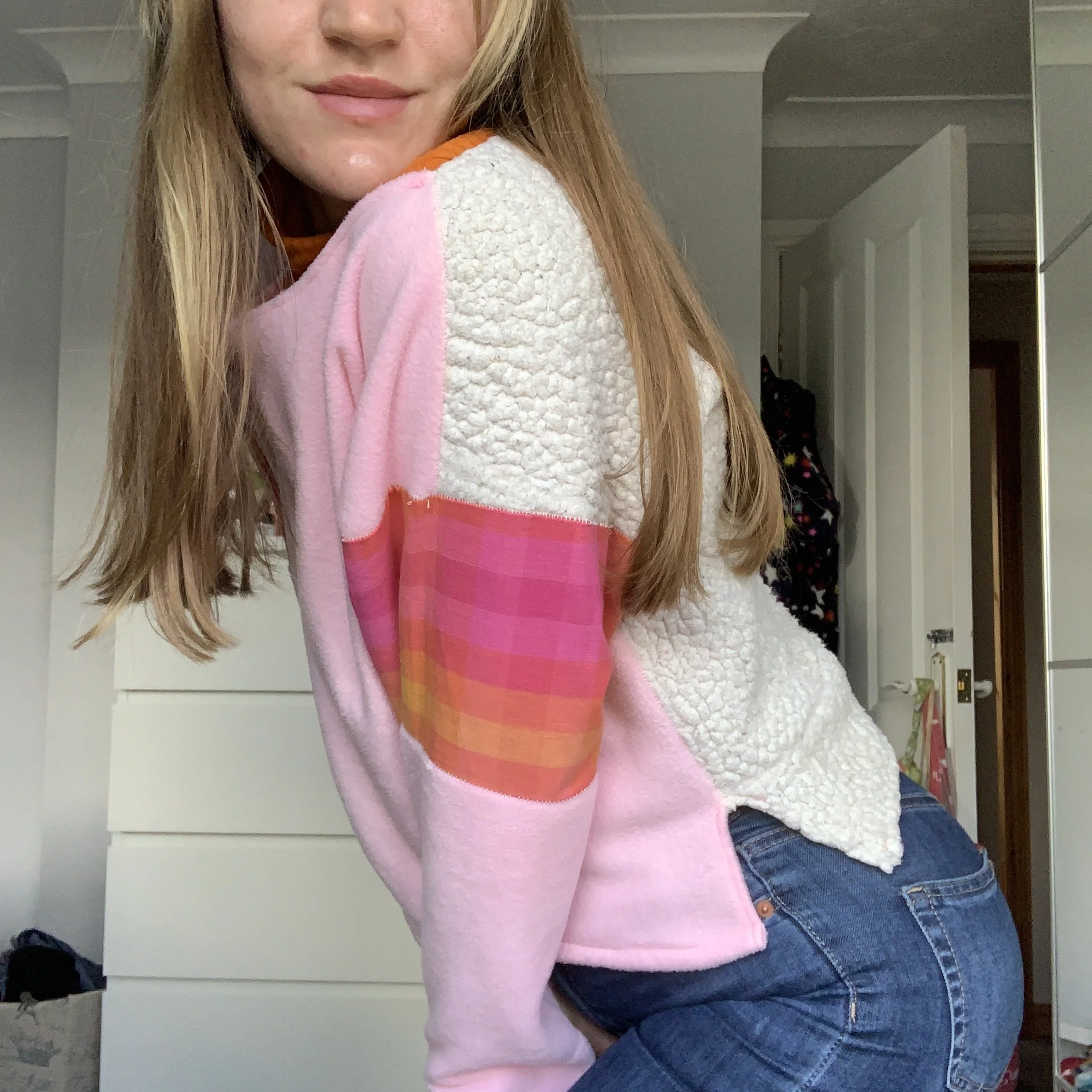 Partial front view of a person with long light brown hair wearing a jumper with pink fleece front, white fleece back with a pink, orange, and yellow plaid patch on the sleeve to the elbow. and blue jeans, in a room with a white dresser and open door.