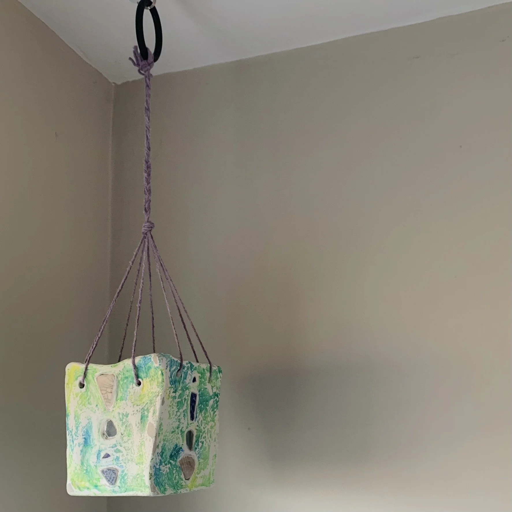 Colorful handmade ceramic pot hanging from a black rubber ring on the ceiling with purple hand dyed string. The pot has shells and sea glass inlaid into the surface.