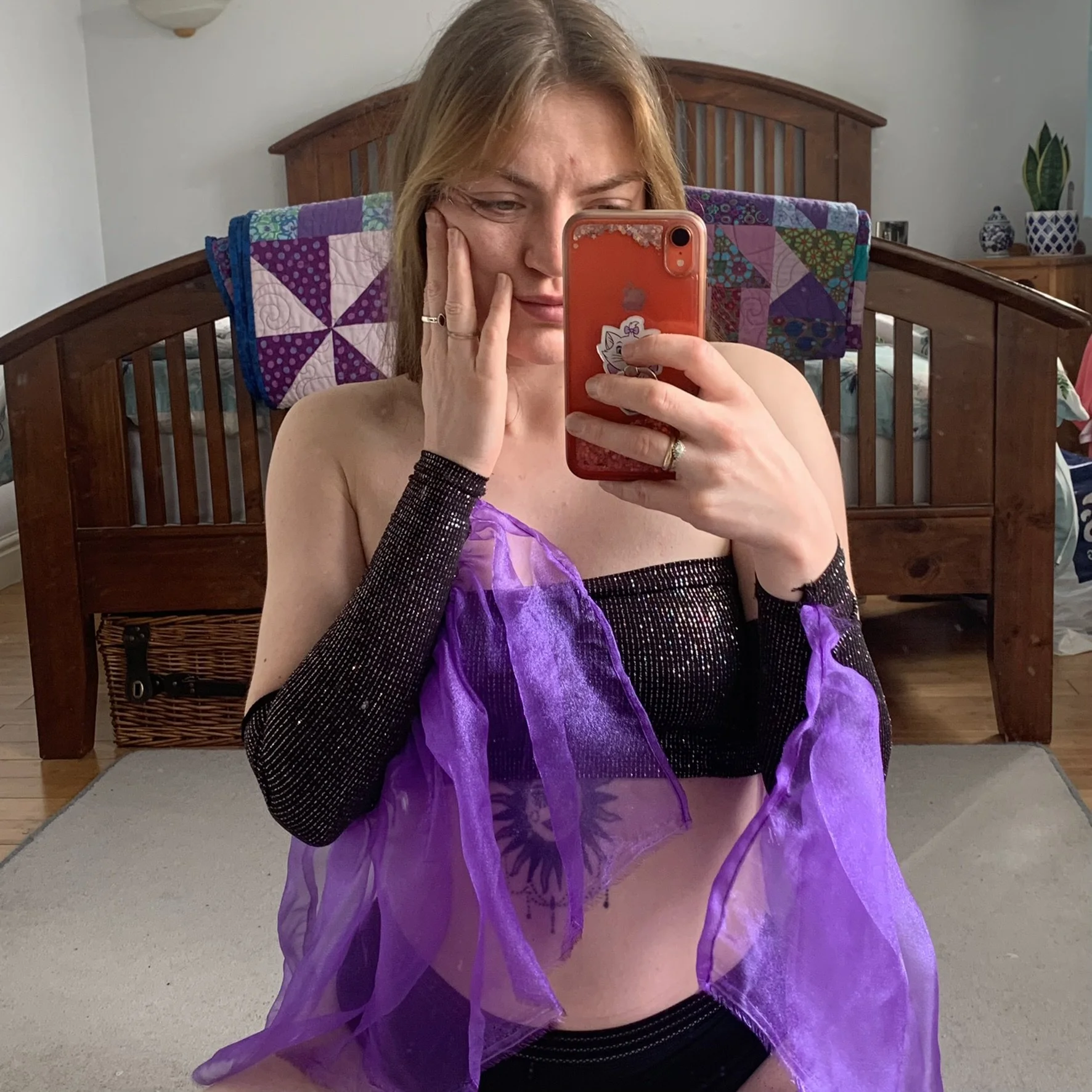 A person taking a mirror selfie in a bedroom. She is wearing black sparkly sleeves and a boob tube with purple mesh flowing from the sleeves of the gloves, with a quilt hanging over a wooden headboard behind her.