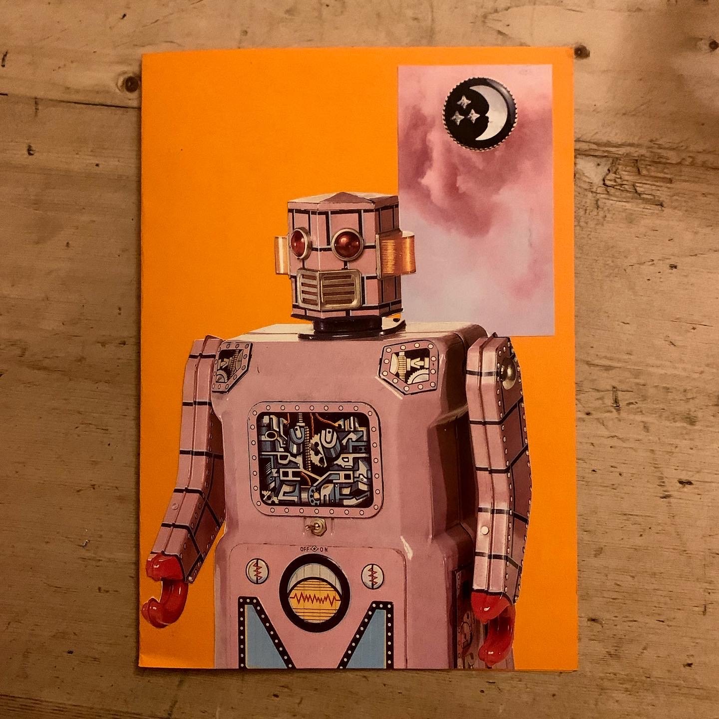 A robot with a pink body, mechanical details, and red hands, standing against an orange background with a small inset image of a pink sky and a black moon with stars.