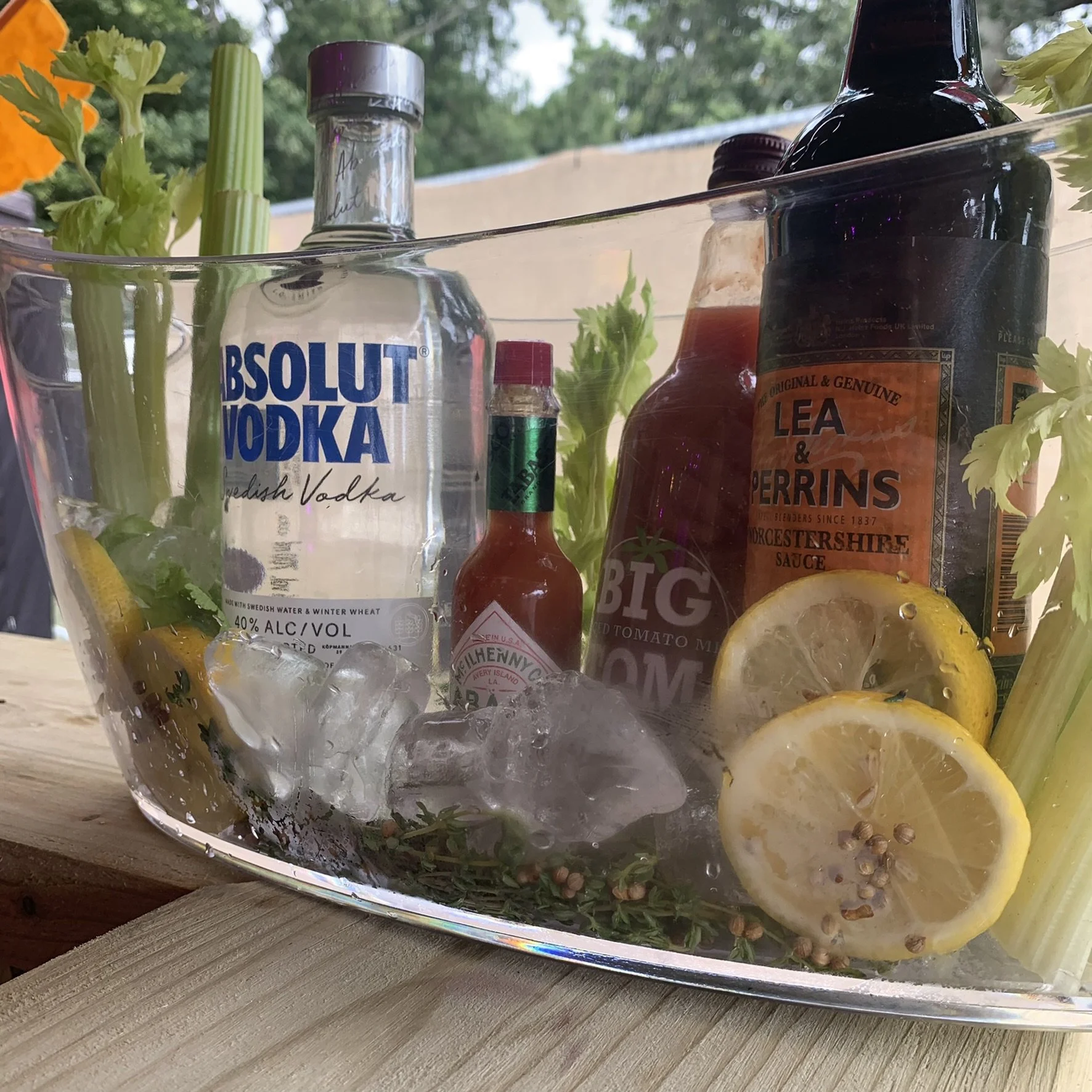 A glass container filled with ice, lemon slices, celery, thyme, and various bottles including Absolut vodka, sriracha sauce, Worcestershire sauce, and hot sauce, with a background of greenery. For a bloody mary marketing display.