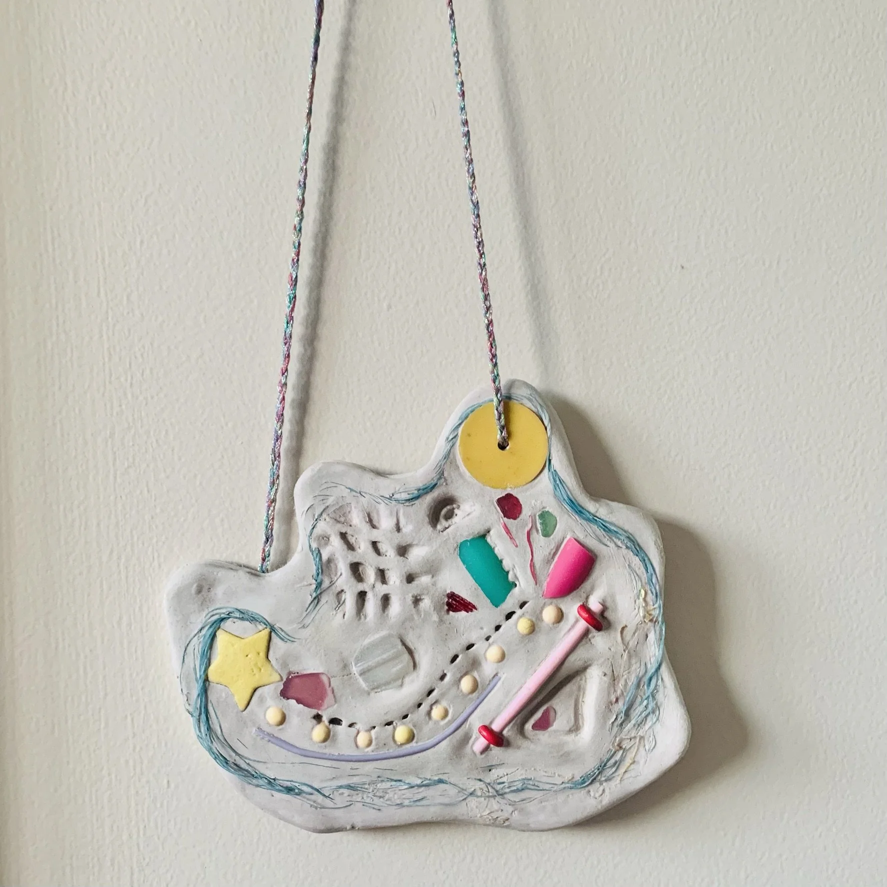Colorful abstract ceramic art piece hanging from a plaited metallic embroidery threads. It features assembled small objects, including stars, dots and circles as well as irregular shapes, with decorative lines and patterns carved into surface.