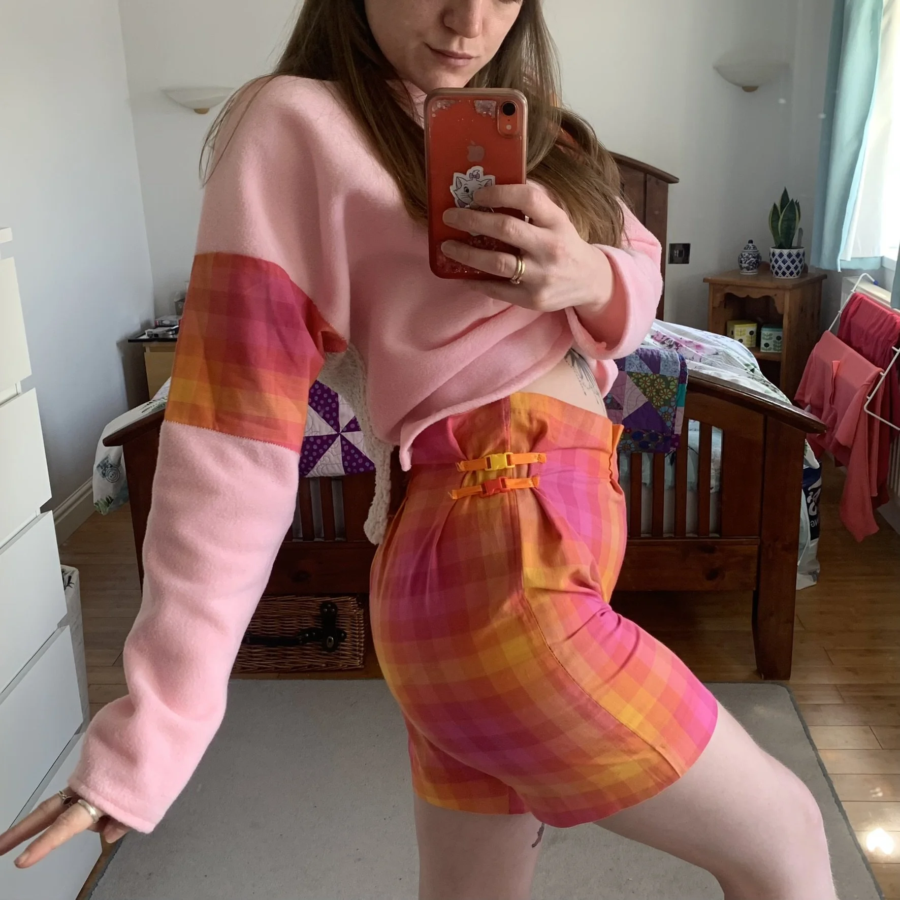 A person taking a mirror selfie, wearing pink and orange plaid shorts with yellow and red buckles and a pink sweatshirt with matching plaid panel sleeves, in a bedroom with a bed, dresser, and laundry drying rack in the background.