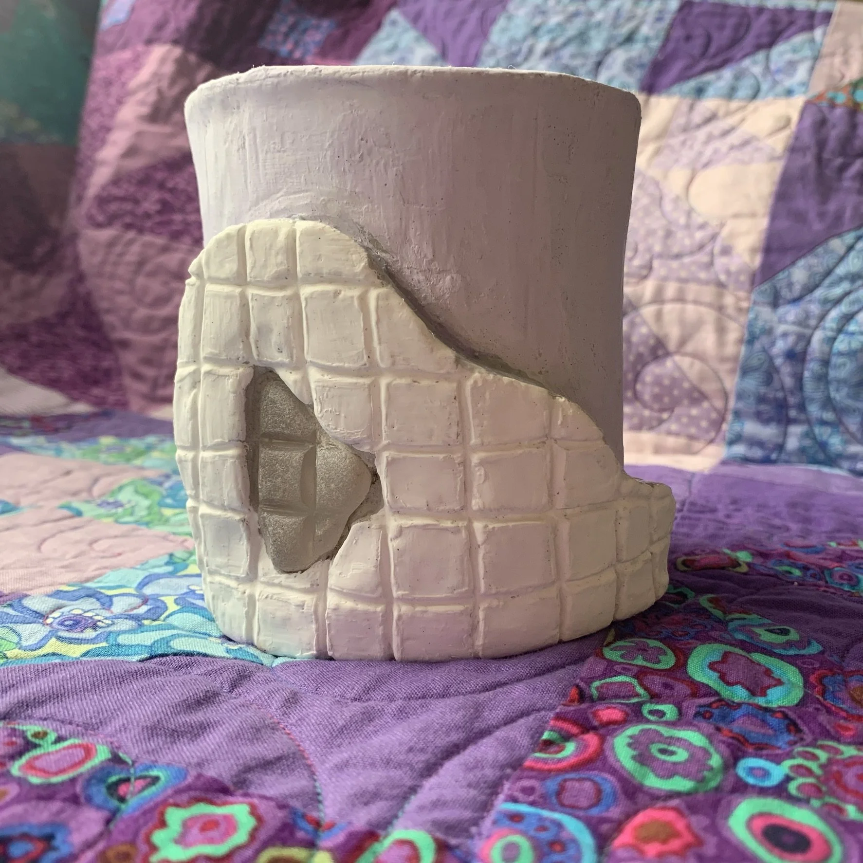 A white and lavender ceramic pot with a textured grid surface, featuring a large, irregular shaped peice of grid textured sea glass inlaid into a raised abstract form on the surface of the pot.