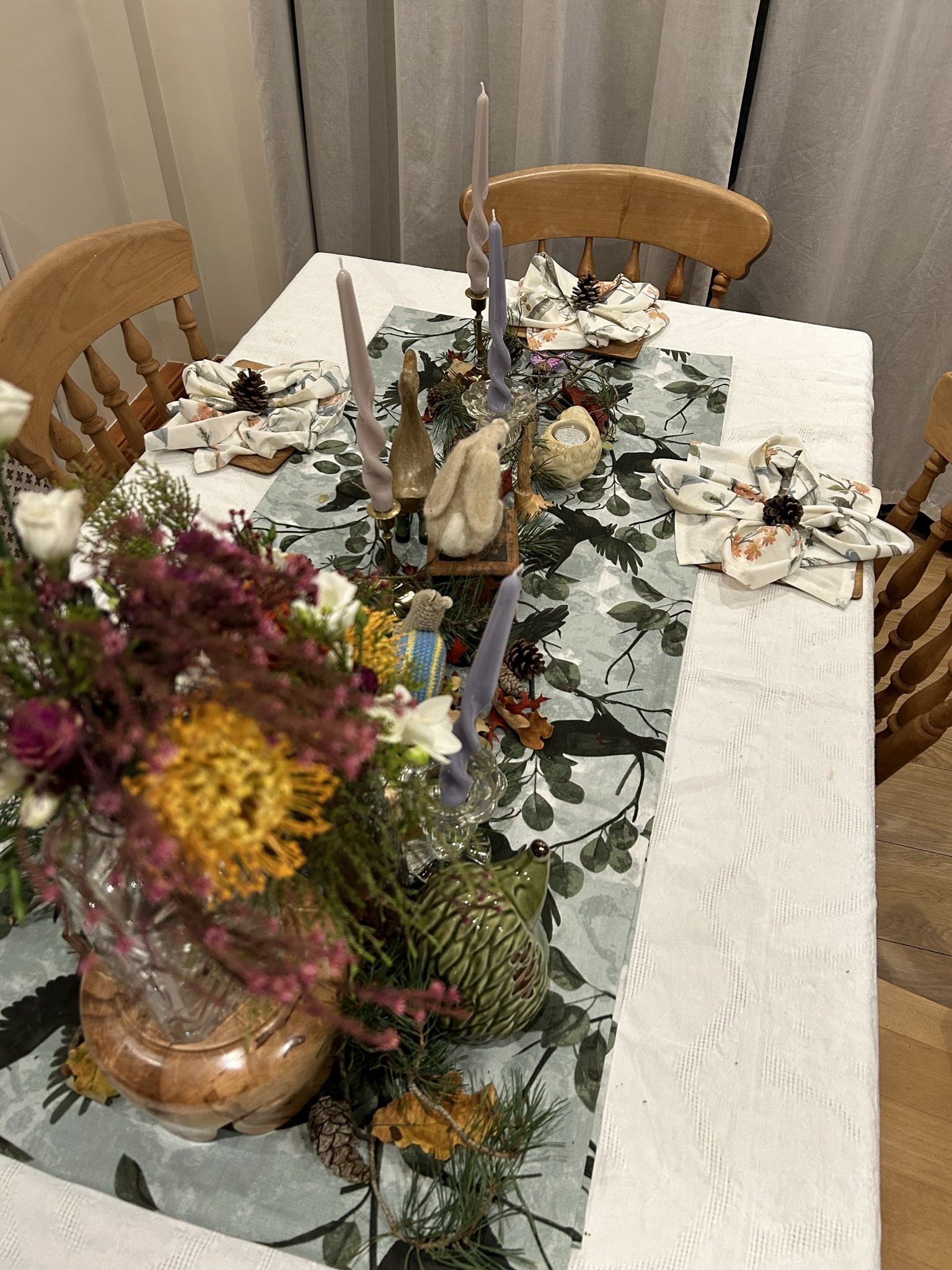 A decorated dining table with a floral table runner, candles, pinecones, and autumn-themed decor, set with four chairs and four napkins.