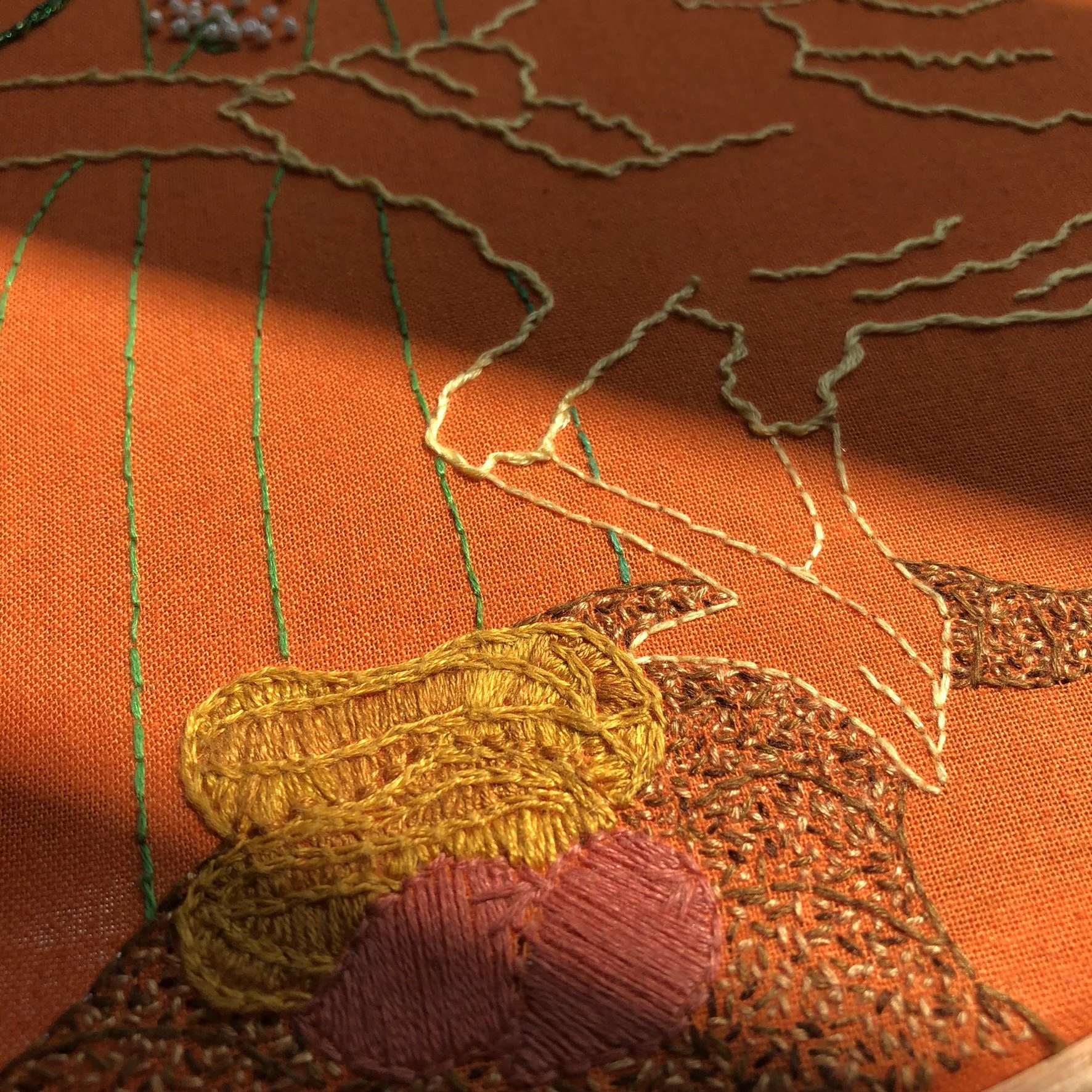 Close-up of an orange fabric with embroidered floral patterns and colorful threads, including yellow, pink, and brown, creating detailed flower designs.