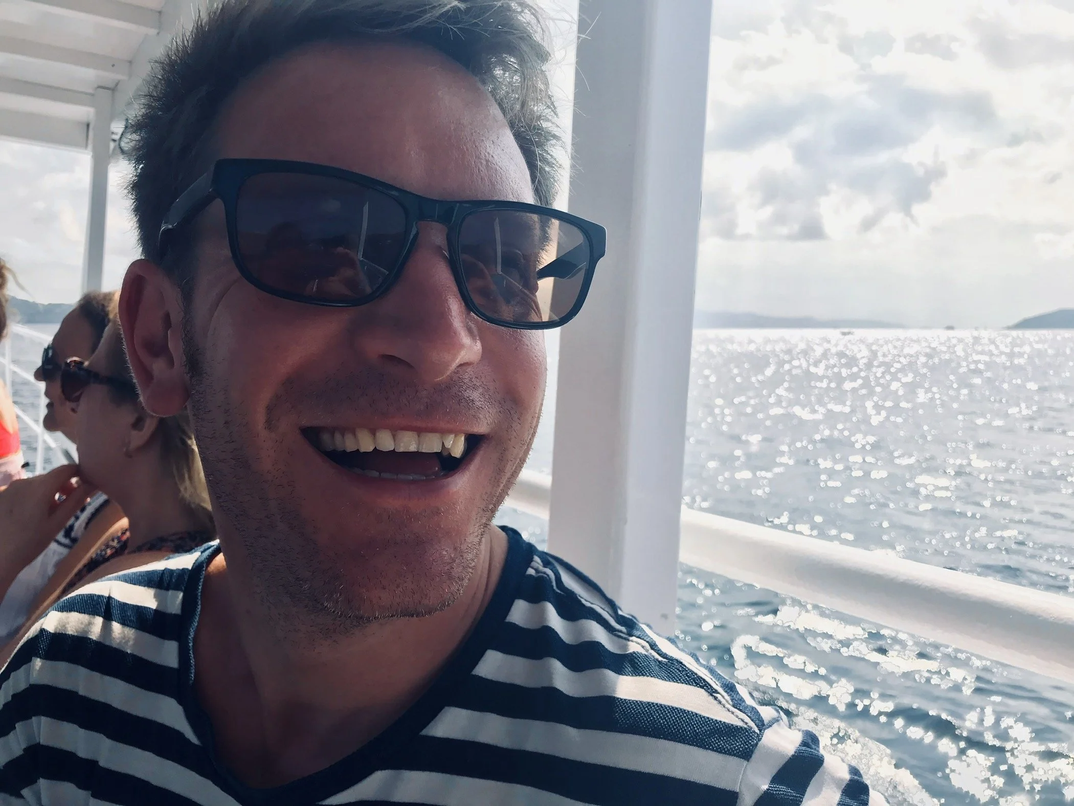 A man wearing sunglasses and a striped shirt smiling on a boat with water and distant land in the background.