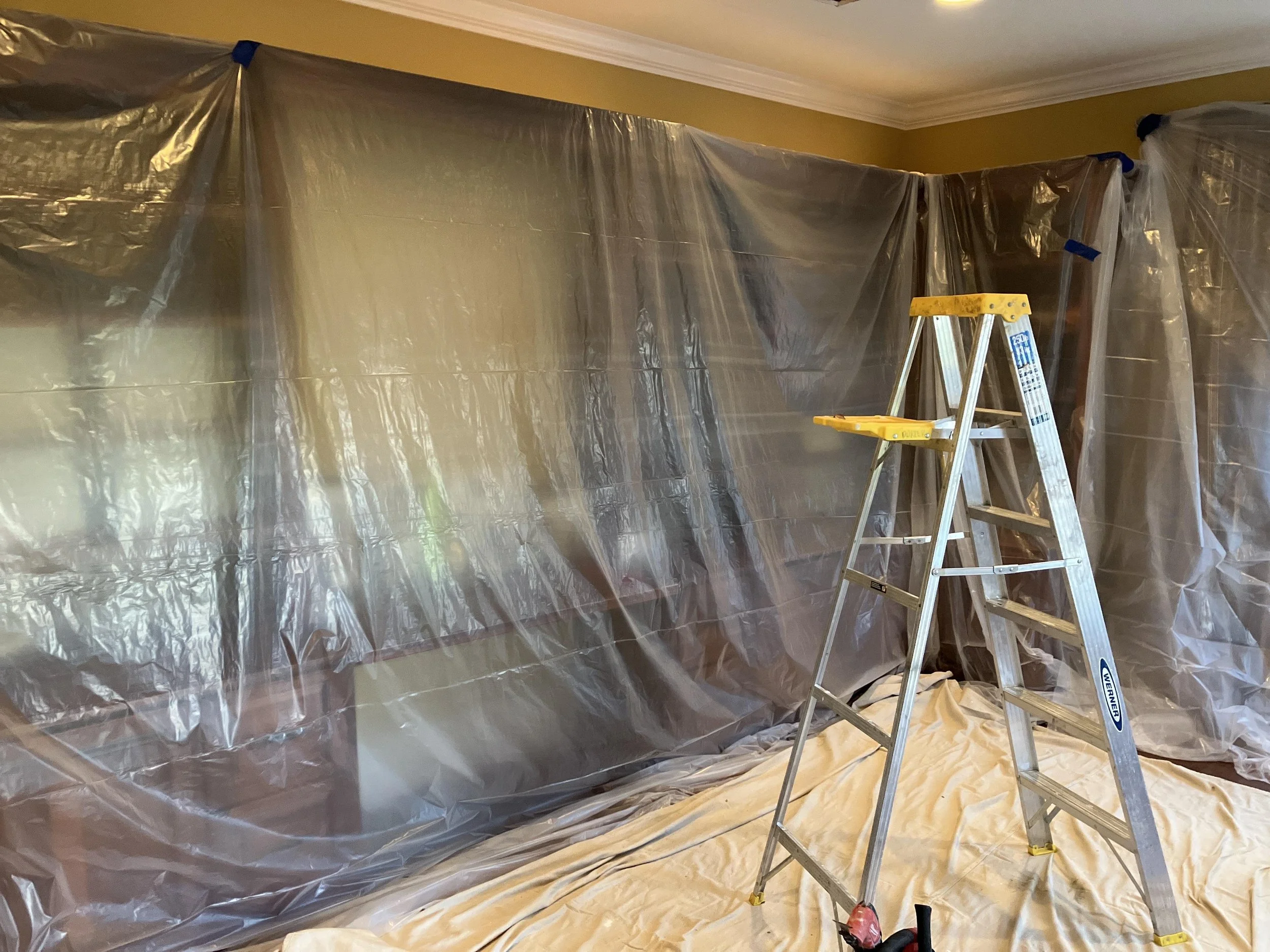 Room covered with large plastic sheeting for painting or renovation. A ladder is positioned in the center, and a drop cloth covers the floor.