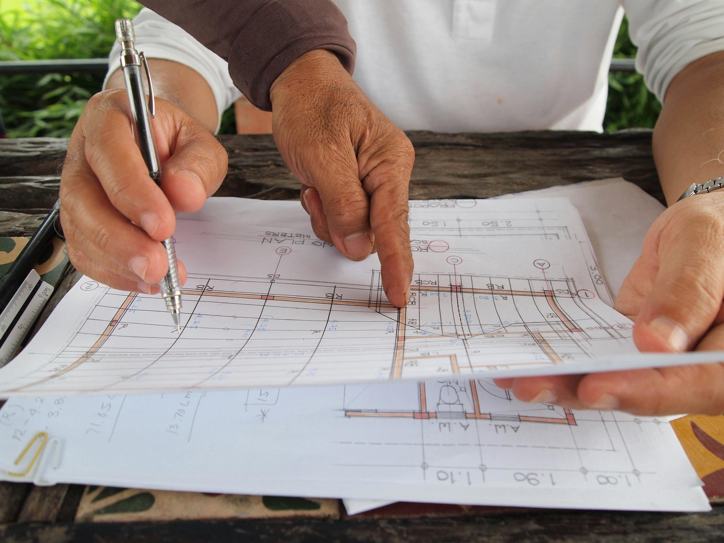 Person pointing at architectural blueprint with a pen, sitting at a rustic outdoor wooden table.