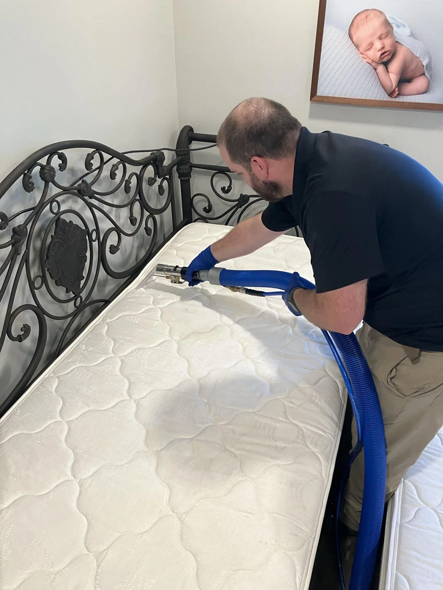 A man cleaning a mattress with a steam cleaner in a bedroom, with a metal bed frame and a framed photo of a sleeping baby on the wall.