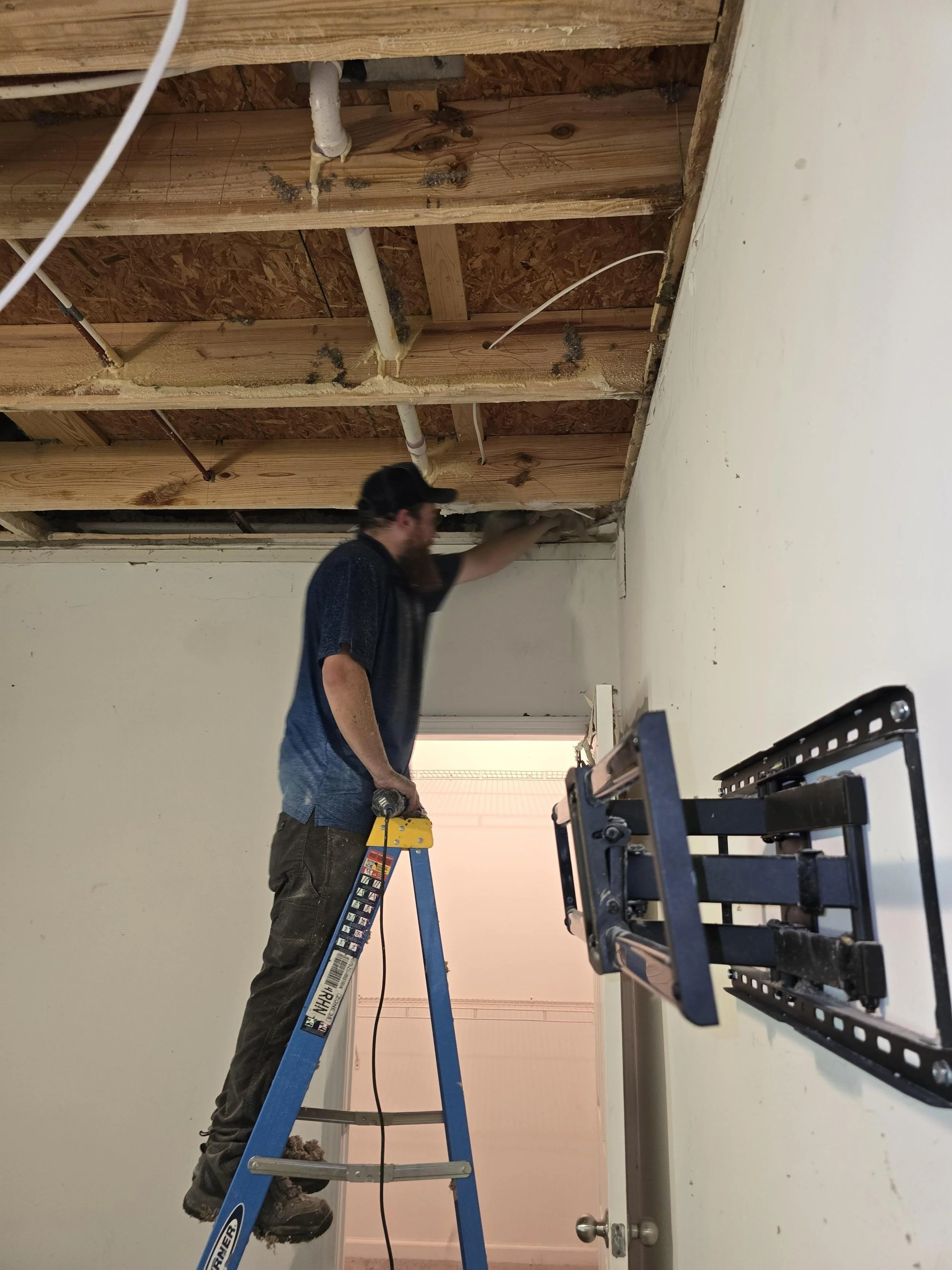 A man standing on a ladder working on the ceiling of a room under construction, with exposed wooden beams and plumbing pipes.