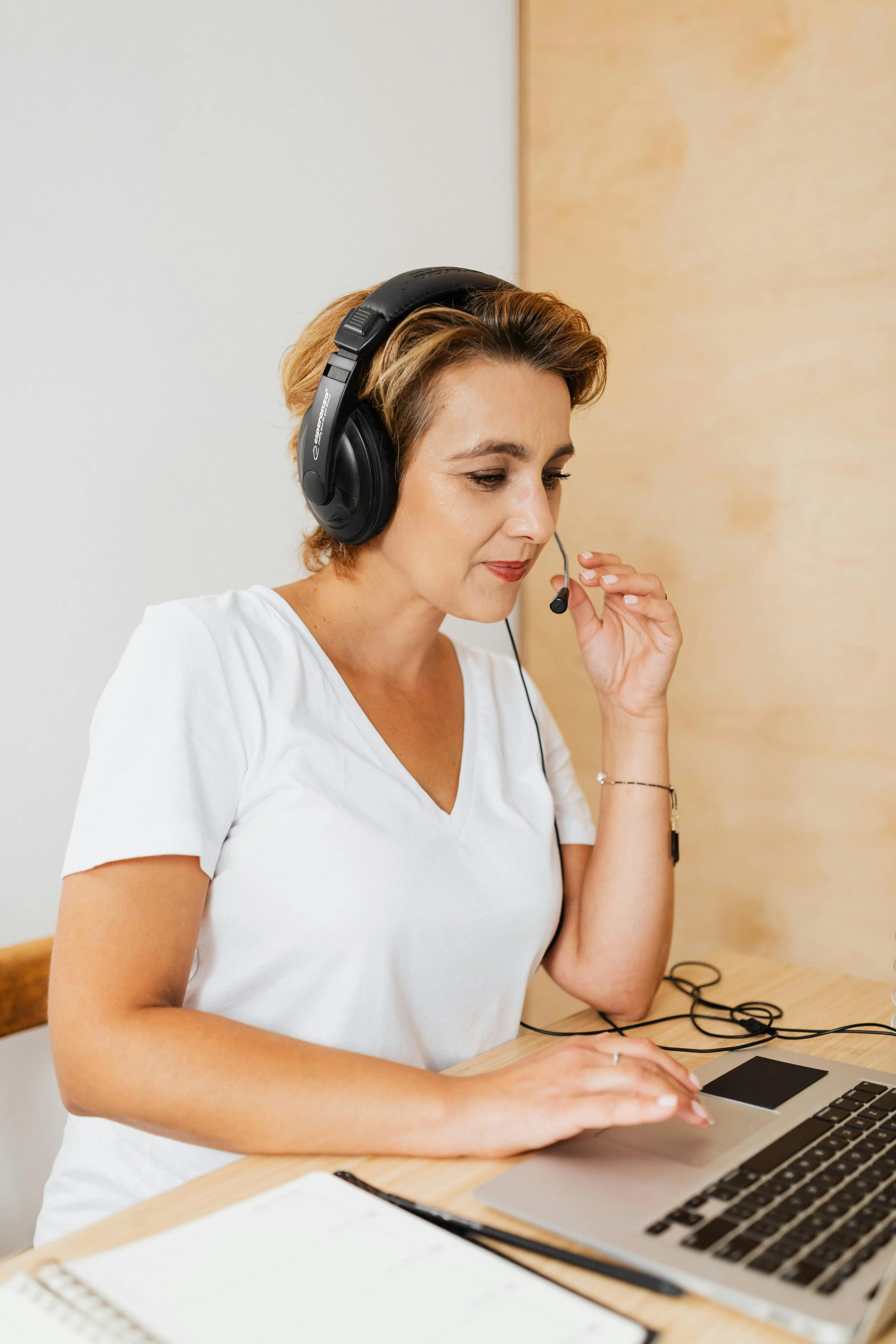 A woman with short light brown hair wearing a white t-shirt and a black headset with a microphone, sitting at a desk with a laptop and open notebooks.