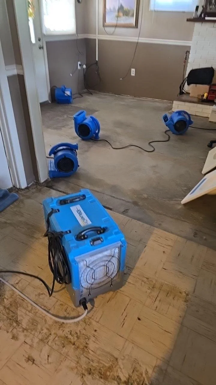 Room with water damage, blue air movers, and a dehumidifier cleaning wet hardwood floors.