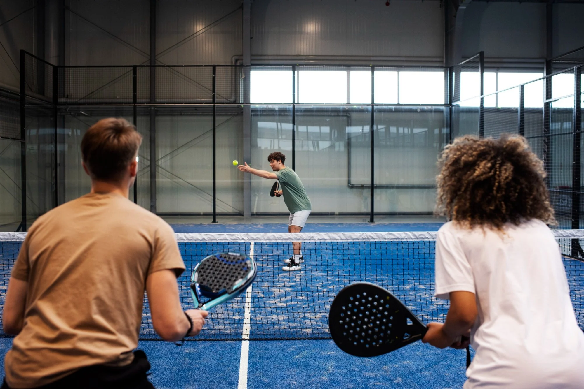 Two people standing on one side of an indoor padel court holding padel rackets, watching a third person serve a green padel ball from the opposite side.