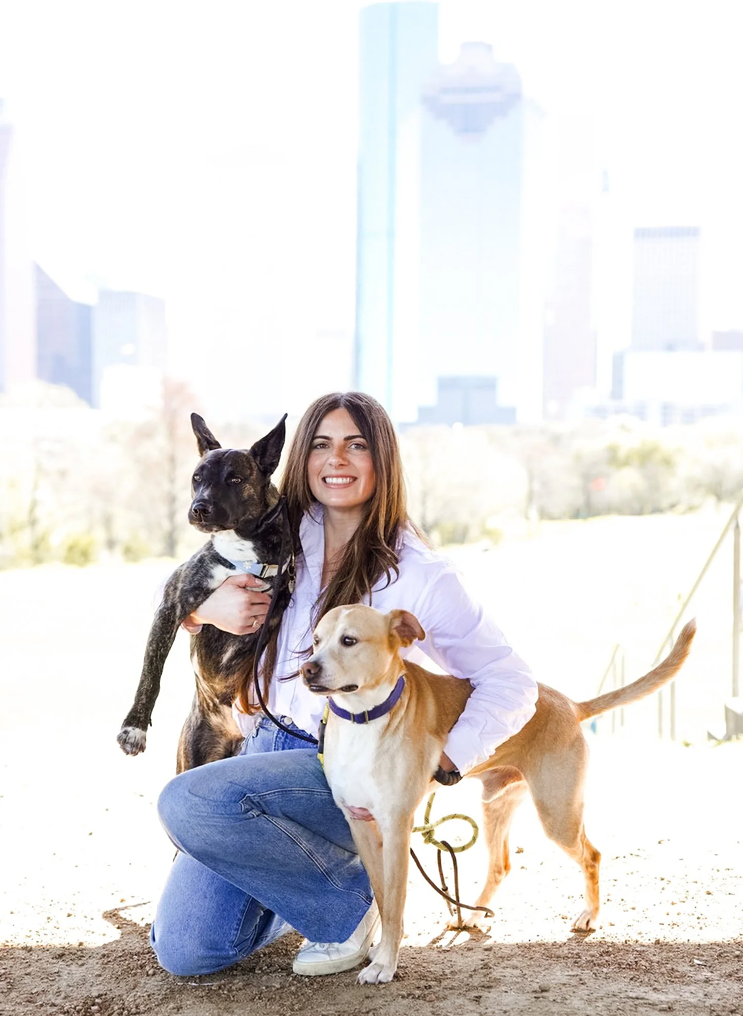 A woman smiling while holding two dogs outside in a park with city buildings in the background.