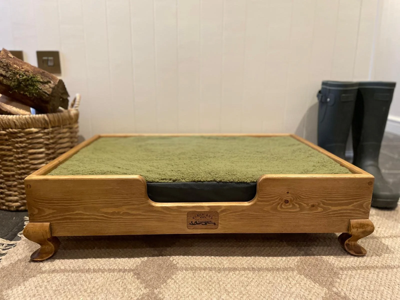 A wooden pet bed with a green cushion inside, situated on a beige carpet. Behind the bed, there is a pair of black rain boots and a basket filled with firewood logs.