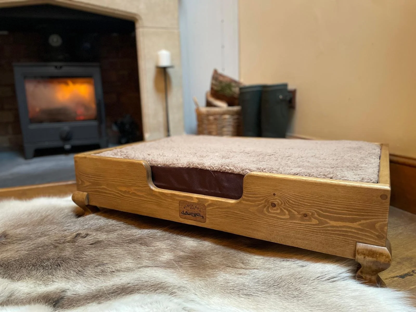 A wooden pet bed with a cushioned top, placed on a animal fur rug in front of a fireplace with a burning fire, and some household items in the background.
