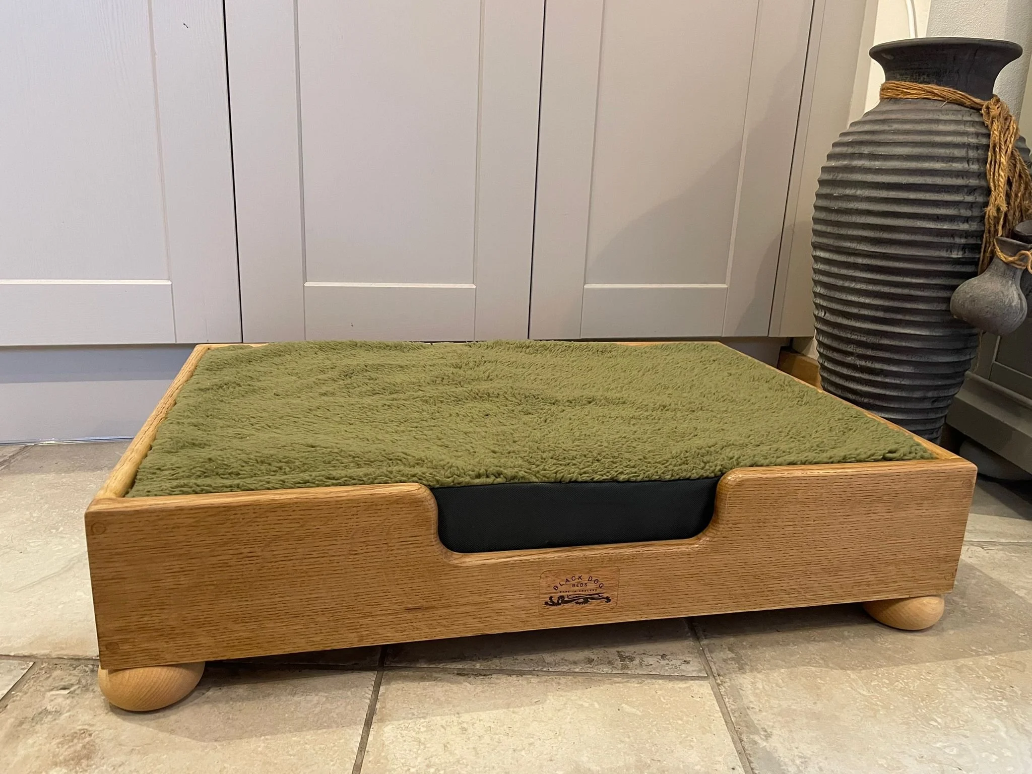 A wooden elevated pet bed with a green fuzzy cushion on top, placed on a tiled floor near a white cabinet and a decorative tall black vase with rope detail.