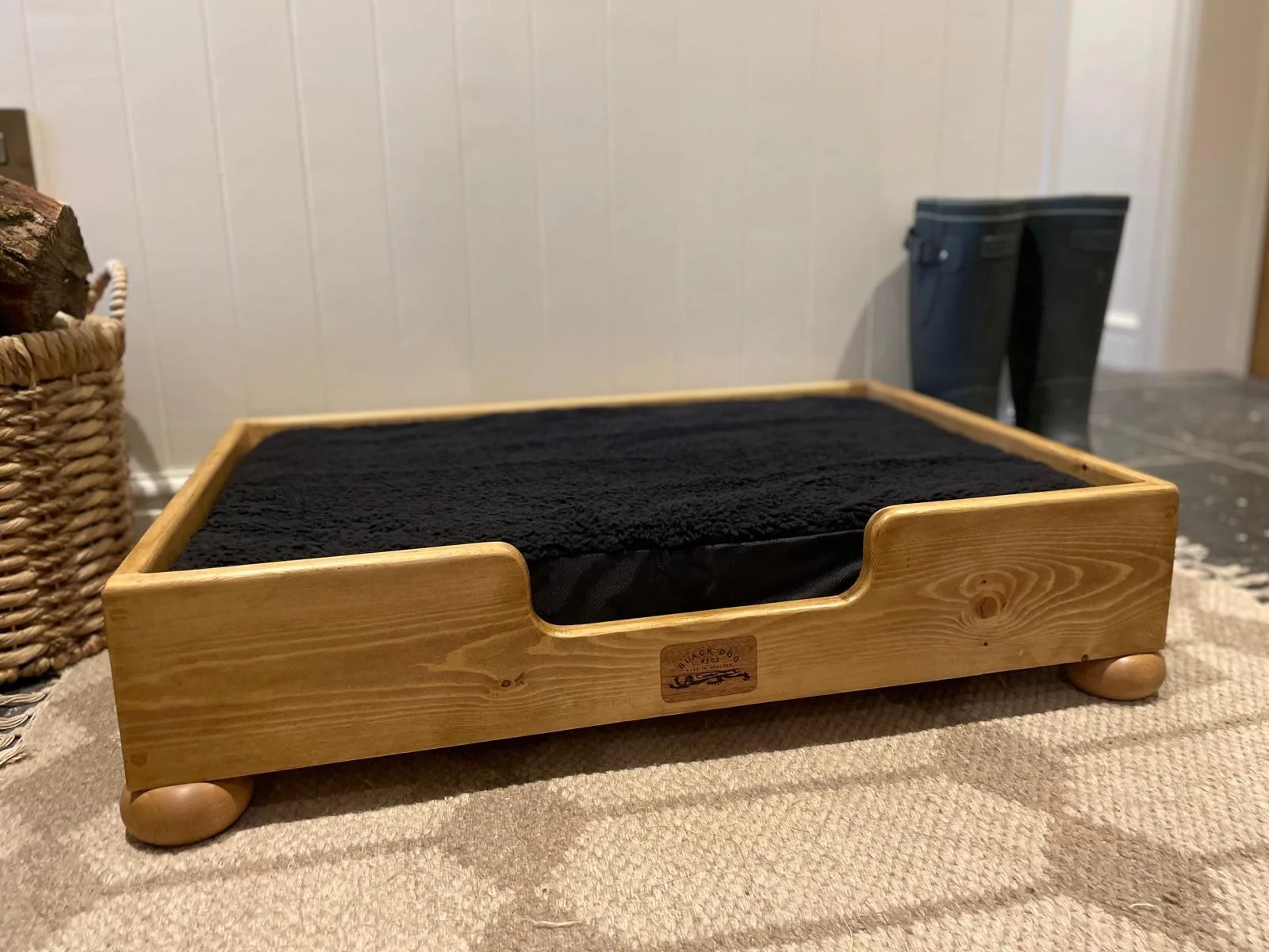 A wooden cat bed with a black cushion on top, placed on a beige textured surface indoors, with a basket and blue rain boots in the background.