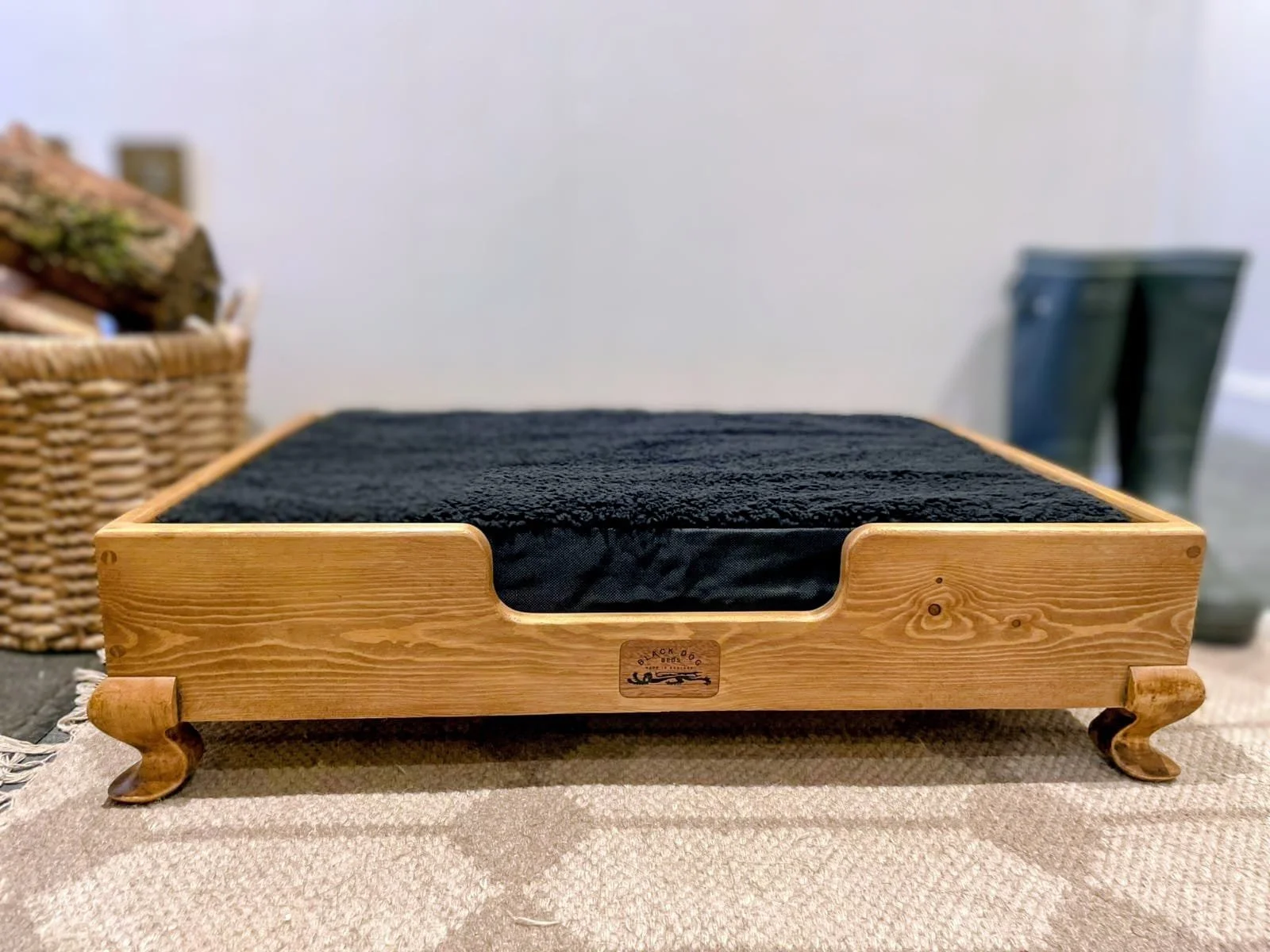 A wooden platform with a black cushion on top, placed on a woven mat. In the background, there is a wicker basket and some objects, with a plain light-colored wall.