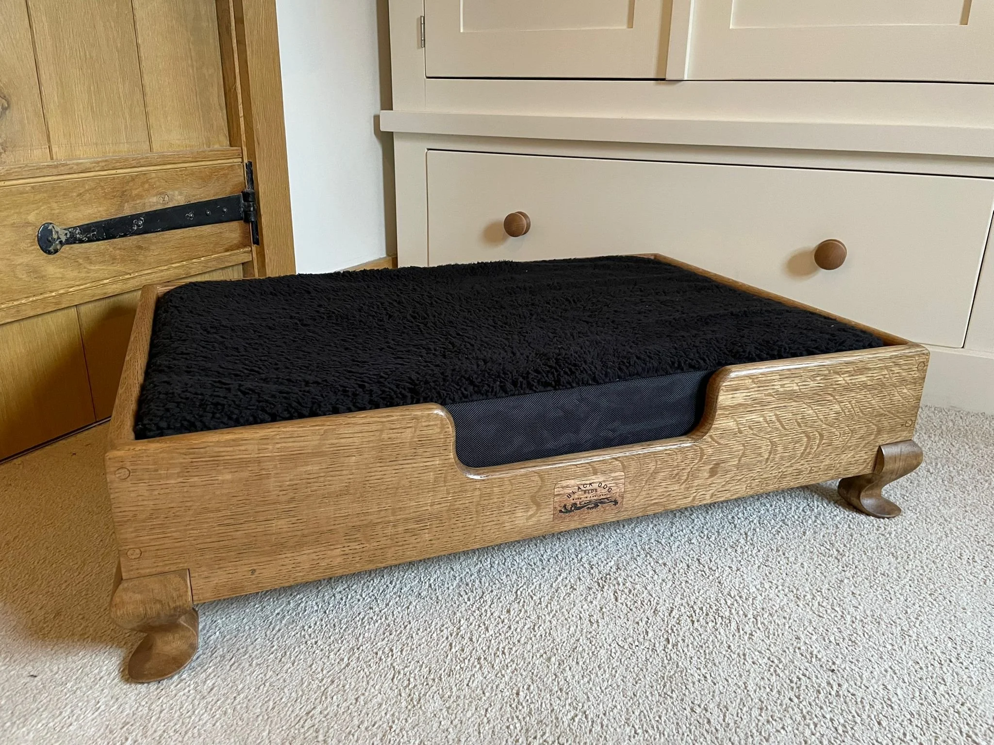 A wooden pet bed with a black cushion inside, placed on beige carpet near a white cabinet and a wooden door.