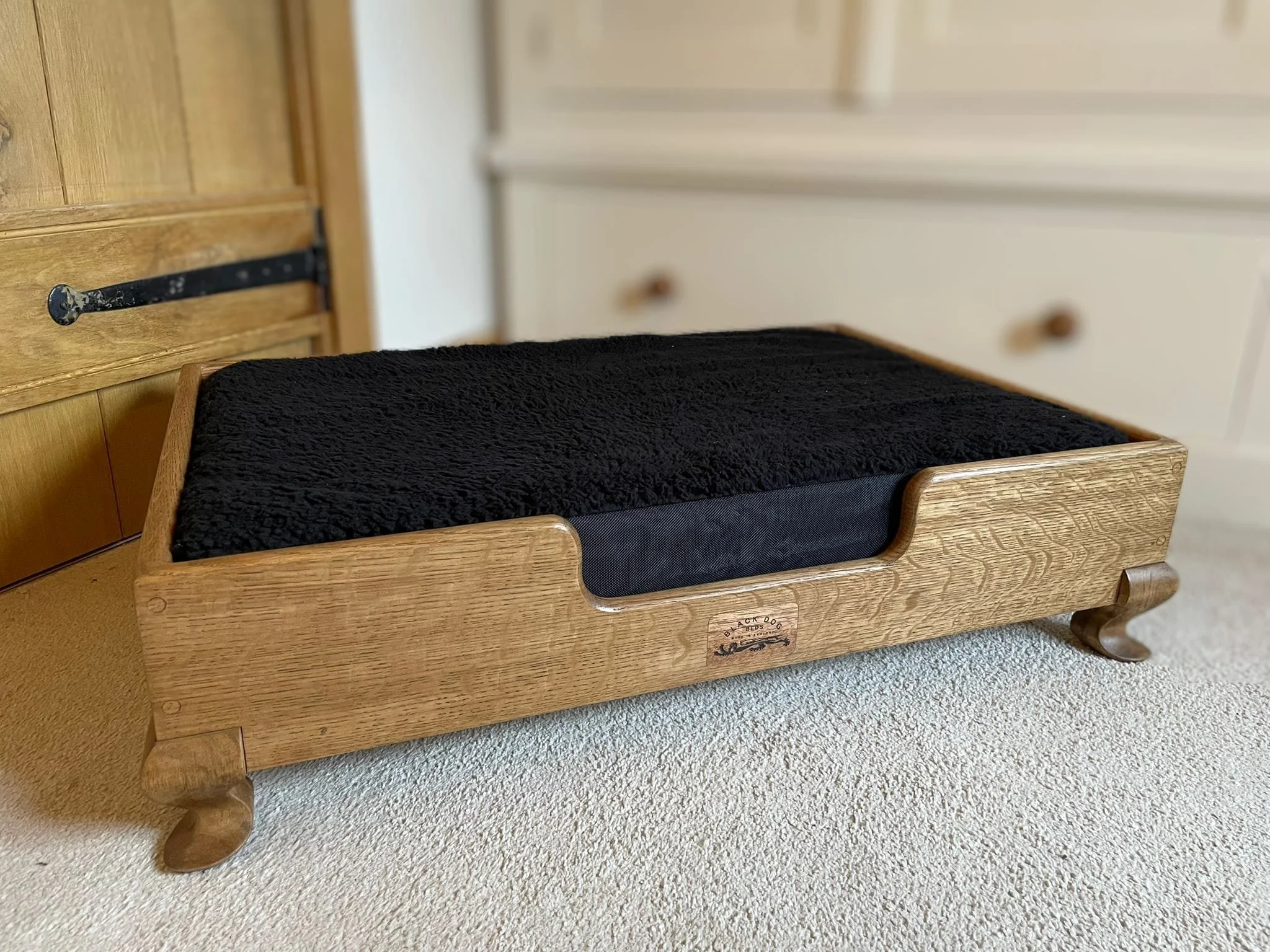 A wooden pet bed with a black cushion on a beige carpet in a room with white walls and wooden furniture.