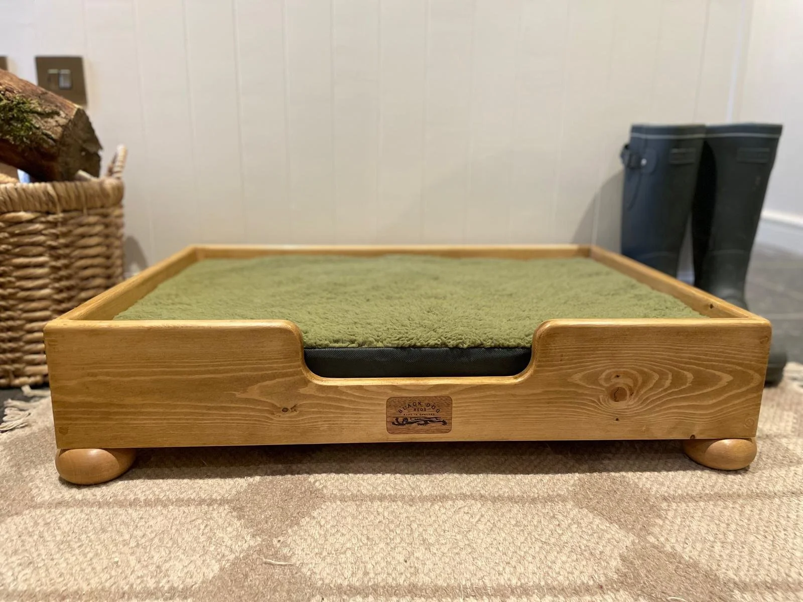 Wooden pet bed with a green cushion, situated on a carpeted floor, with a woven basket and black boots in the background.