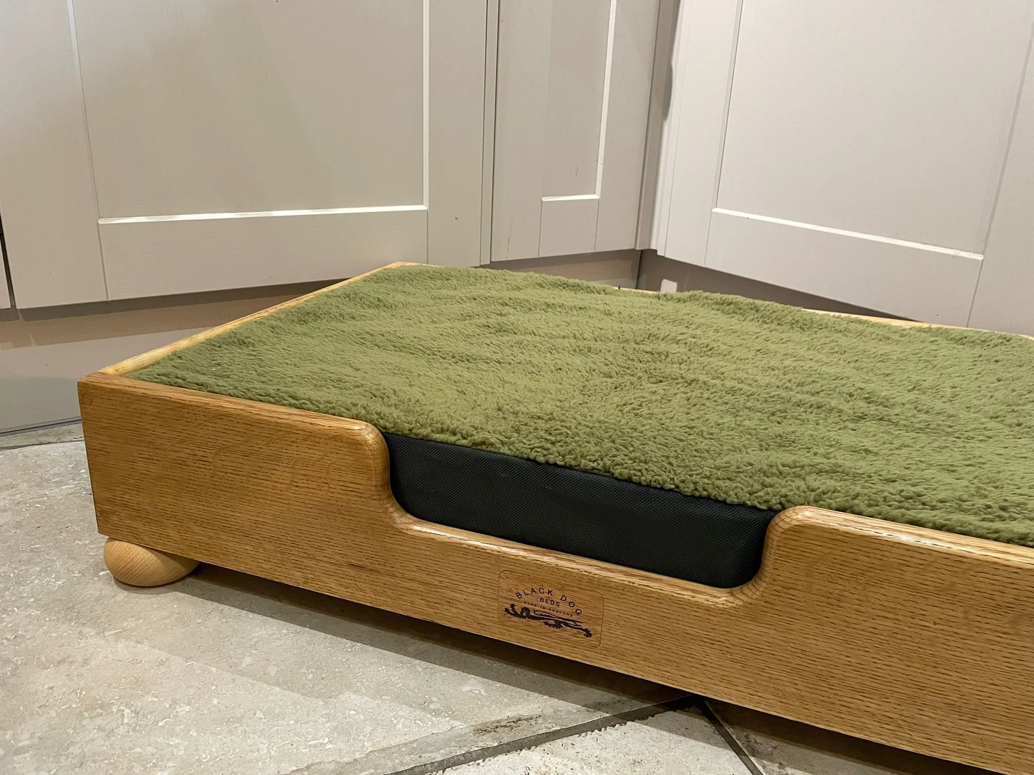 A small wooden bed with a green blanket on a tiled floor in front of white kitchen cabinets.