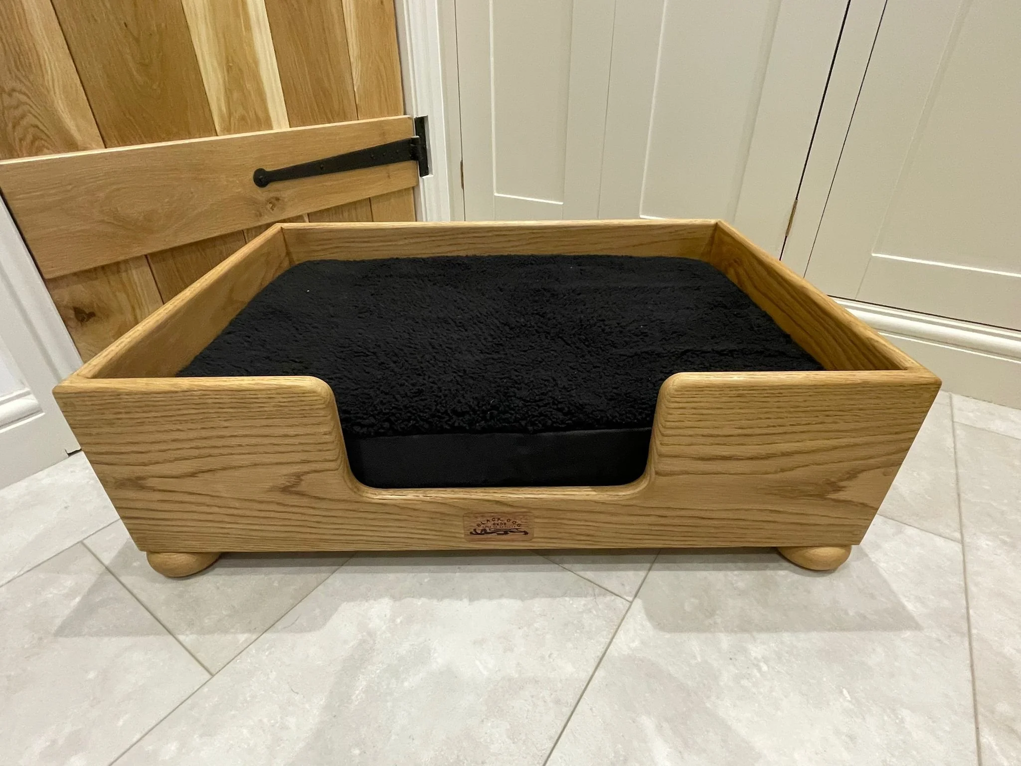 A wooden pet bed with a black cushion inside, placed on a tiled floor in a room with wooden and white cabinets.