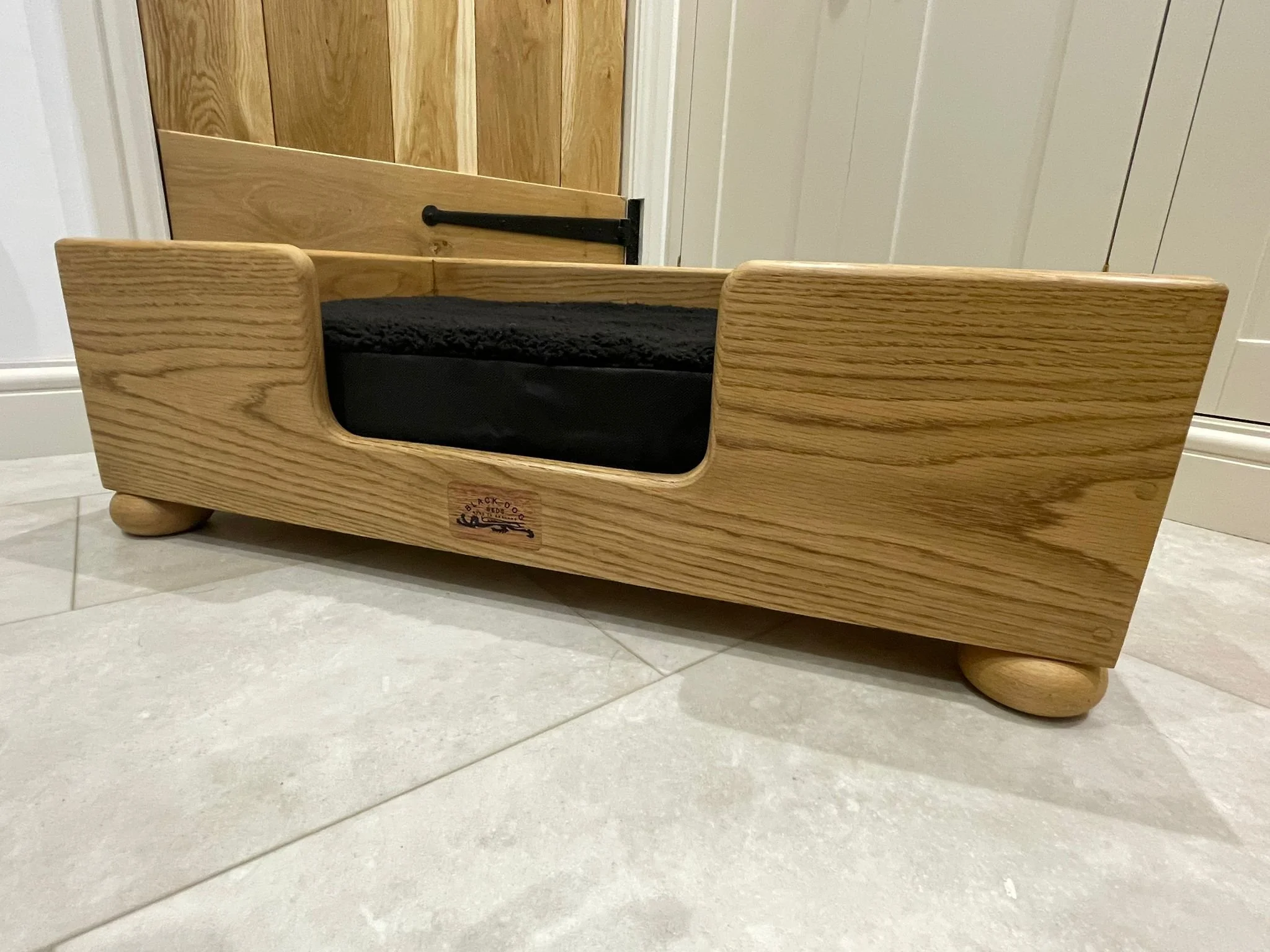 A wooden bed with rounded edges and a black cushion on top, placed on a tiled floor in front of a wooden door with a black latch.