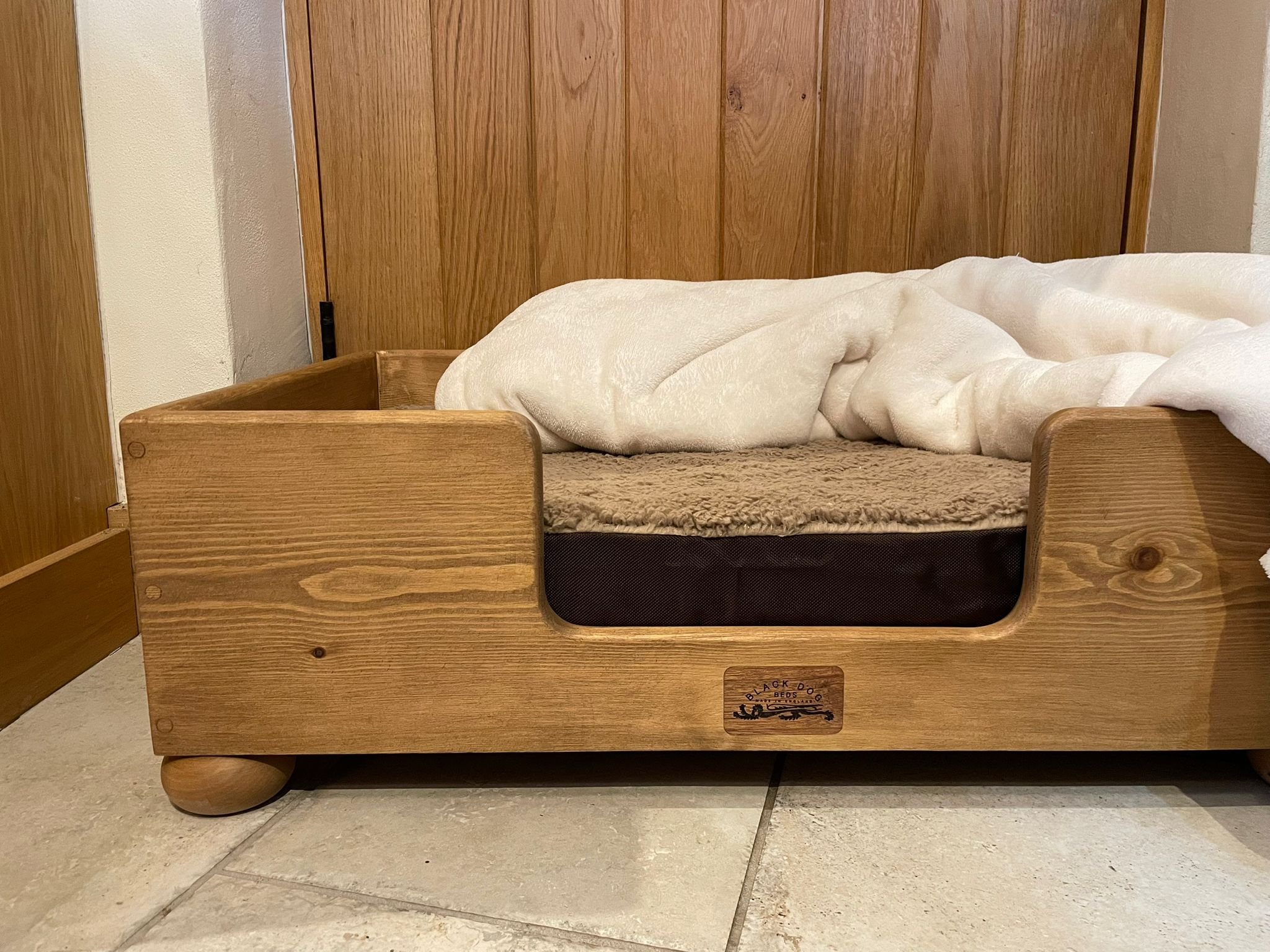 A wooden bed frame with a bed that has a beige blanket and white comforter, positioned on a tiled floor in front of a wooden door.