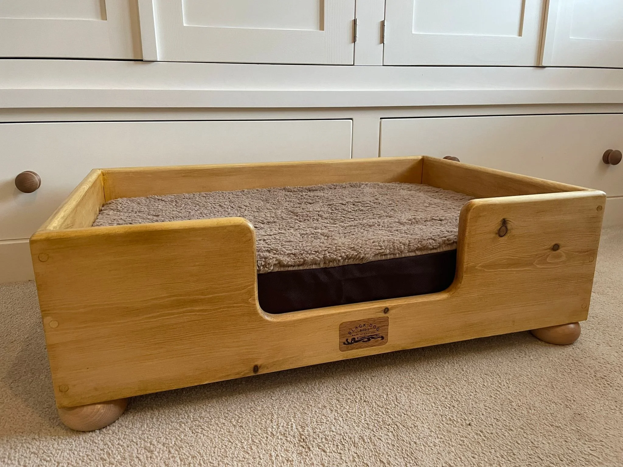 A wooden pet bed with a soft cushion on a carpeted floor, positioned in front of white cabinets.