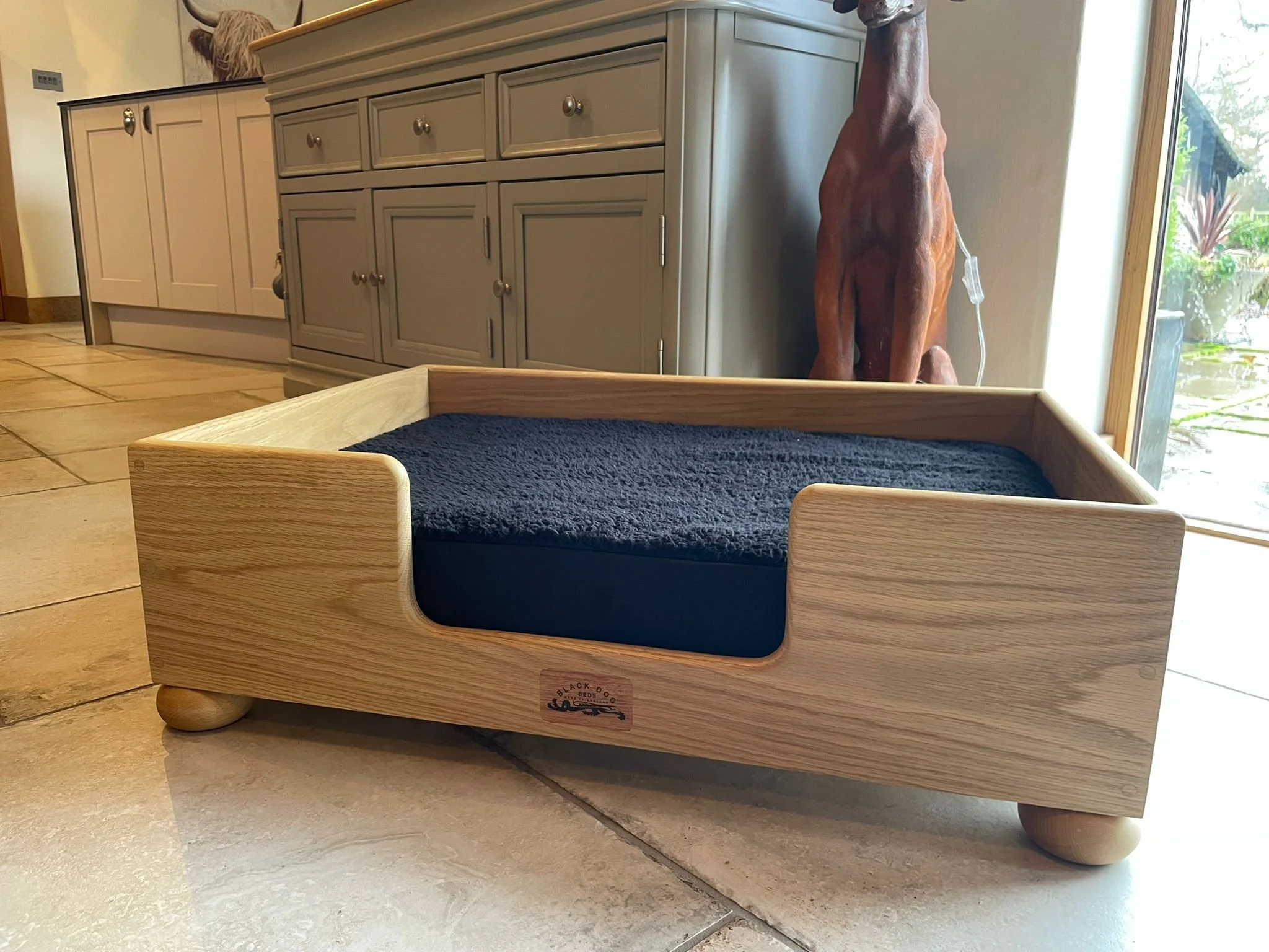 A wooden bed with a black mattress pad, situated near a window with greenery outside, in a room with tile flooring and cabinets.