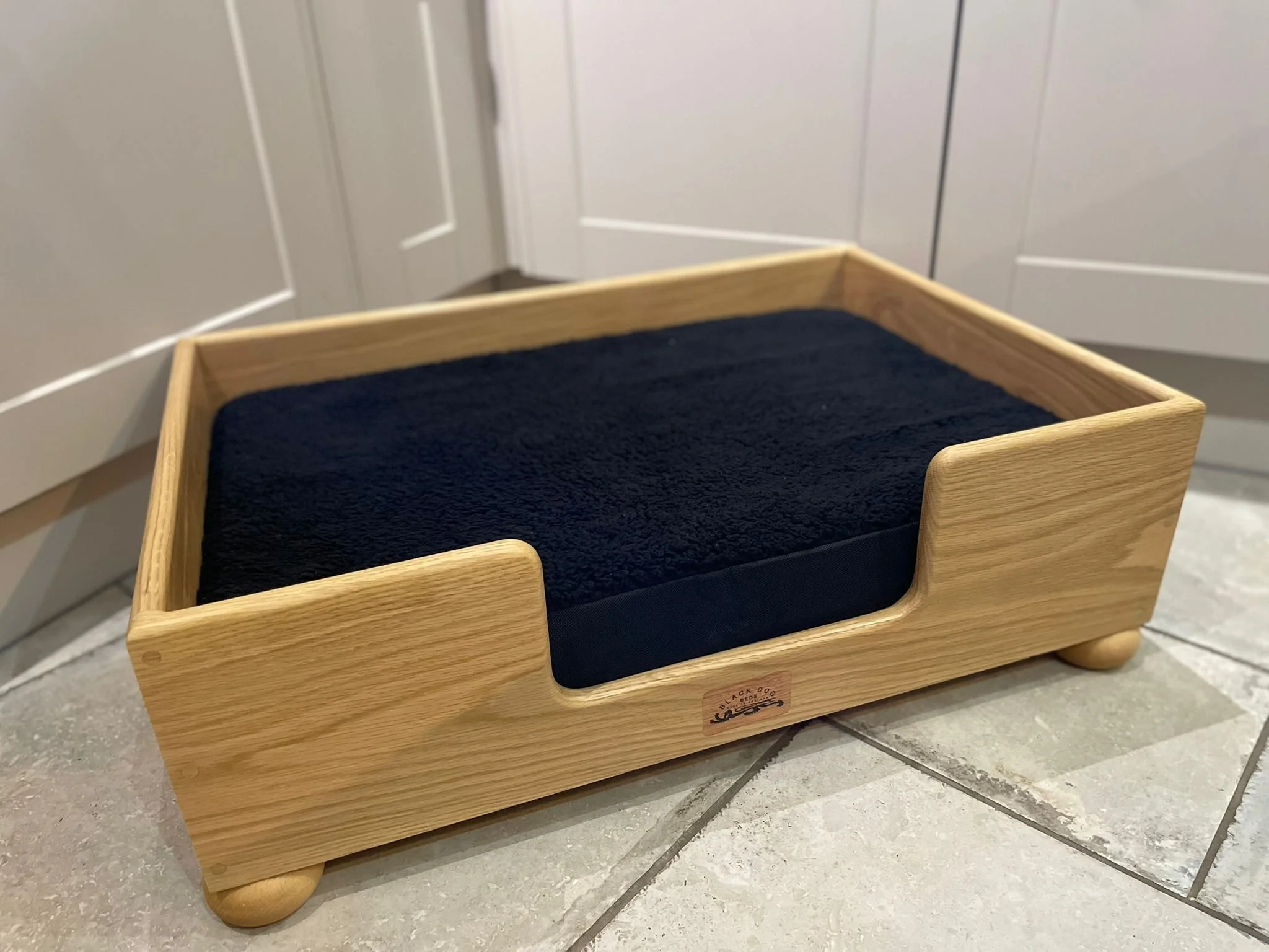 A wooden pet bed with a cushion inside, placed on tan tiled floor near white cabinets.