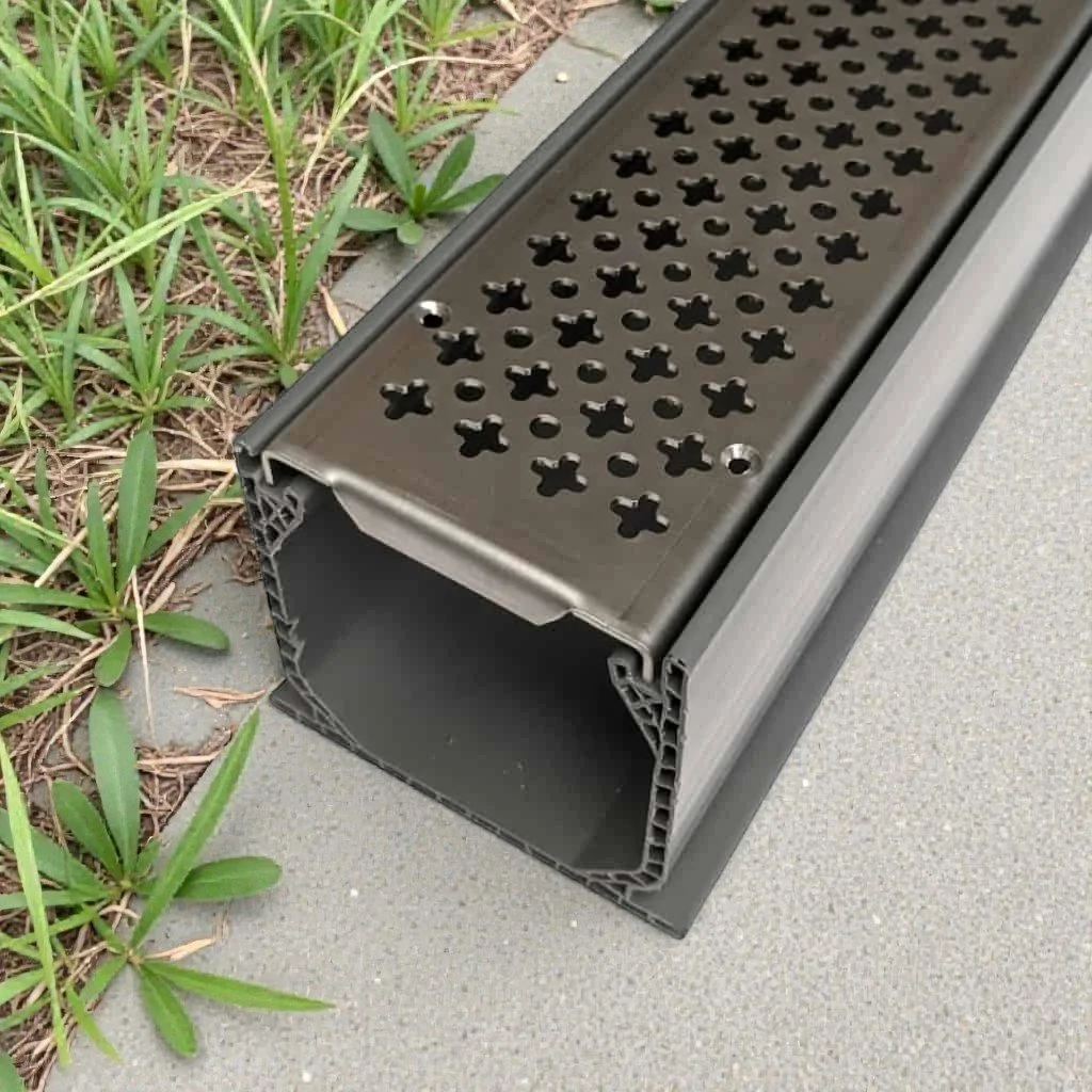 Close-up of a gray metal drainage grate on a concrete sidewalk, with some green grass and plants beside it.