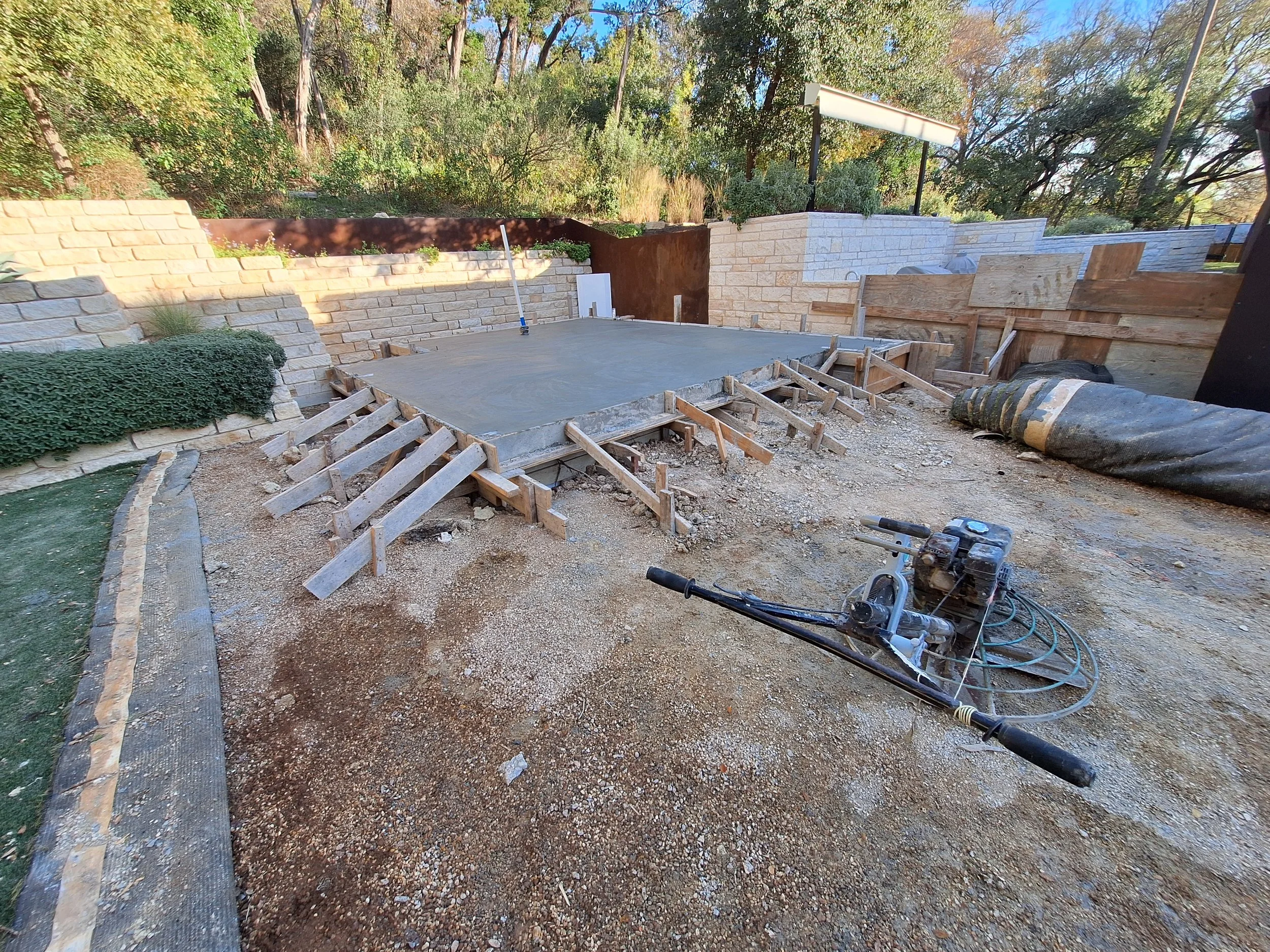 Construction site with a concrete slab foundation, wooden framing, and a power trowel for smoothing concrete, surrounded by gravel and building materials.