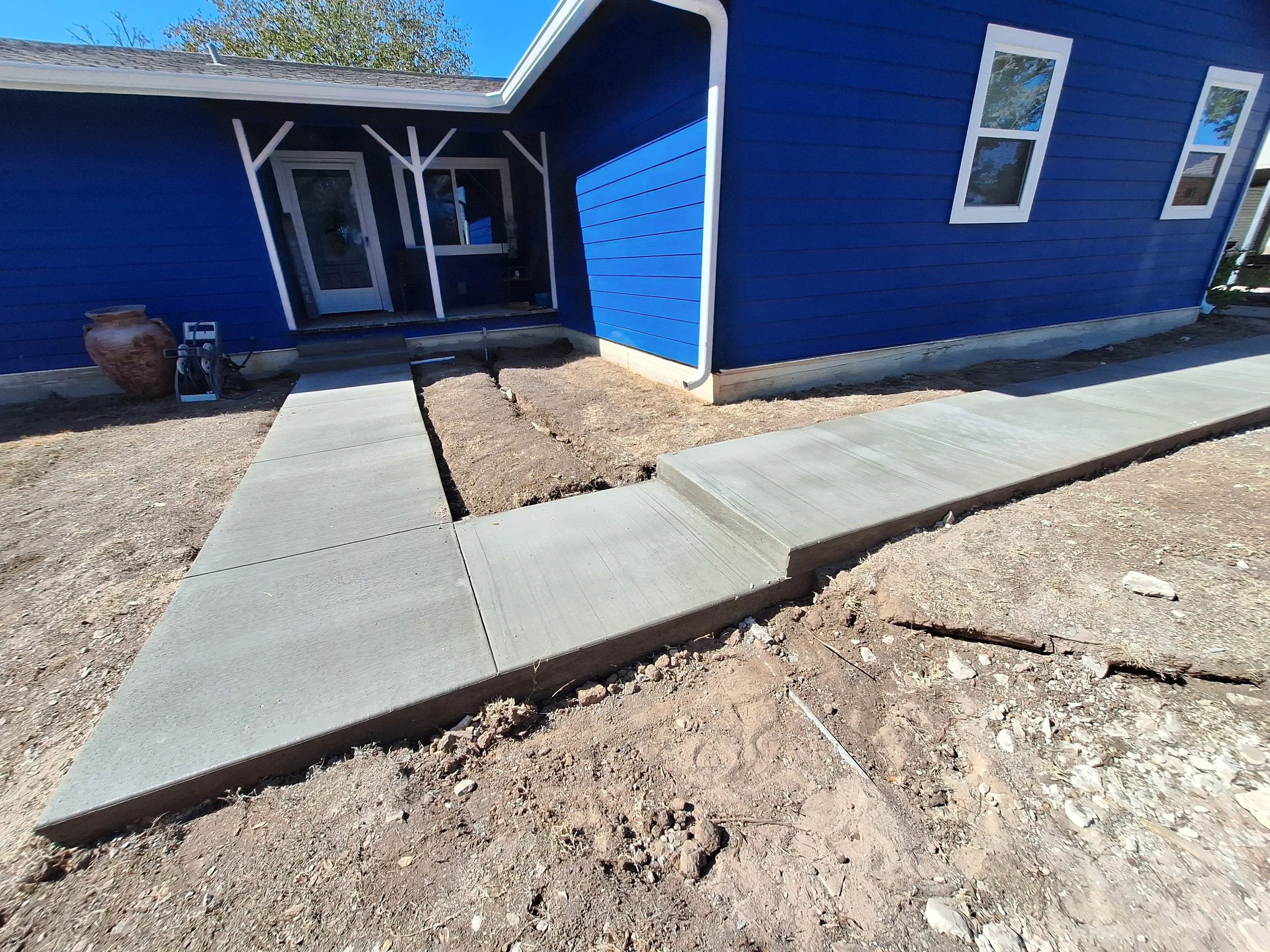 Newly poured concrete walkway leading to the front door of a blue house with white trim, on a sunny day.