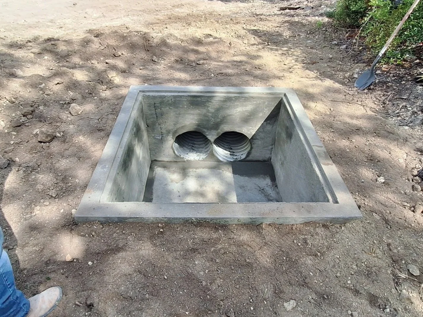 Concrete structure with two circular drain pipes installed at the bottom, set into a dirt ground, with a small shovel leaning against a bush on the right side.