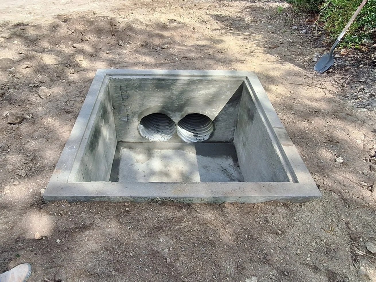 A square concrete structure with two drainage pipes on the side, set into the ground outdoors.