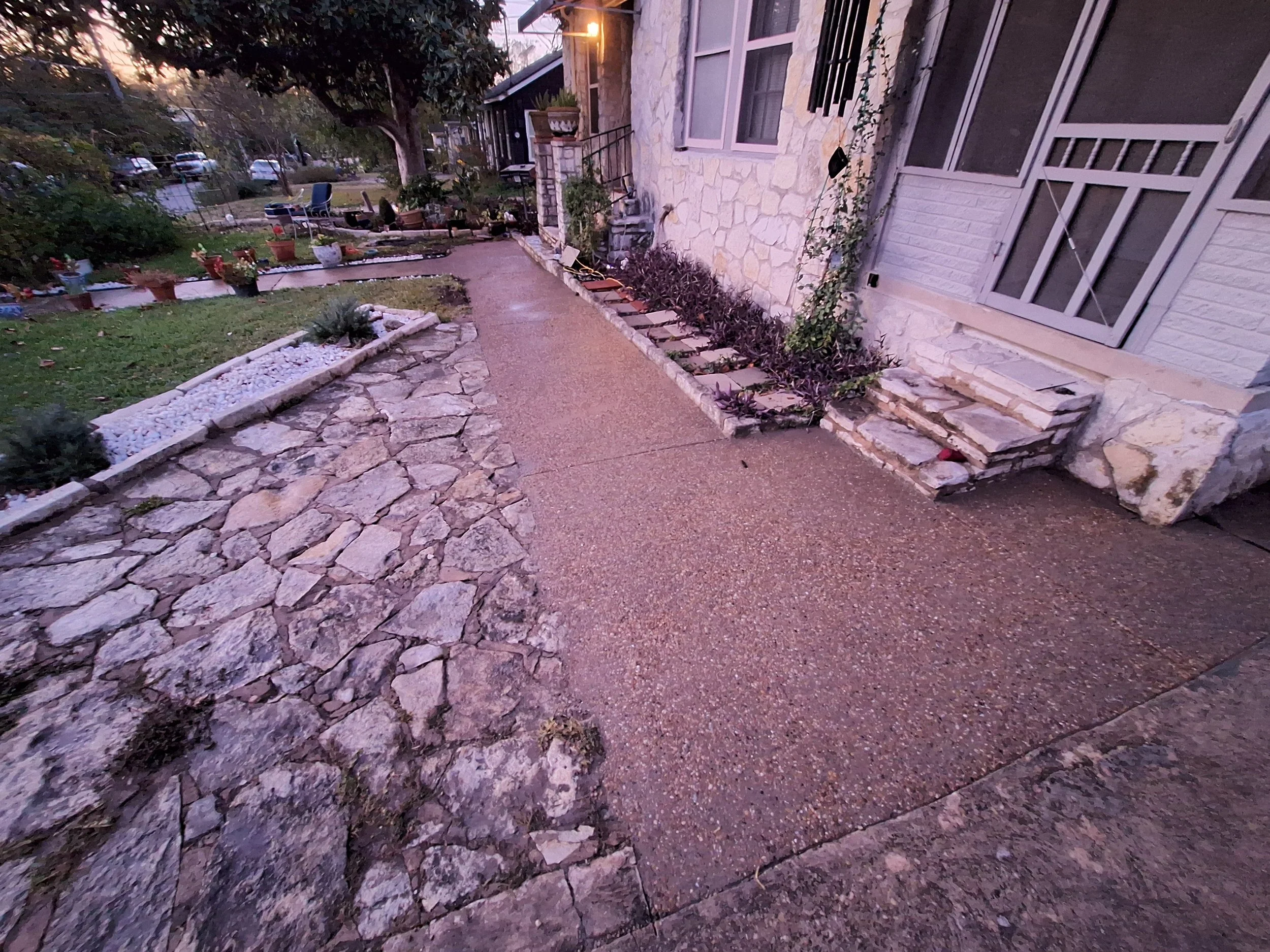A stone pathway in front of a house with similar stone steps leading to the door, surrounded by a garden with potted plants, trees, and bushes, during sunset or dusk.