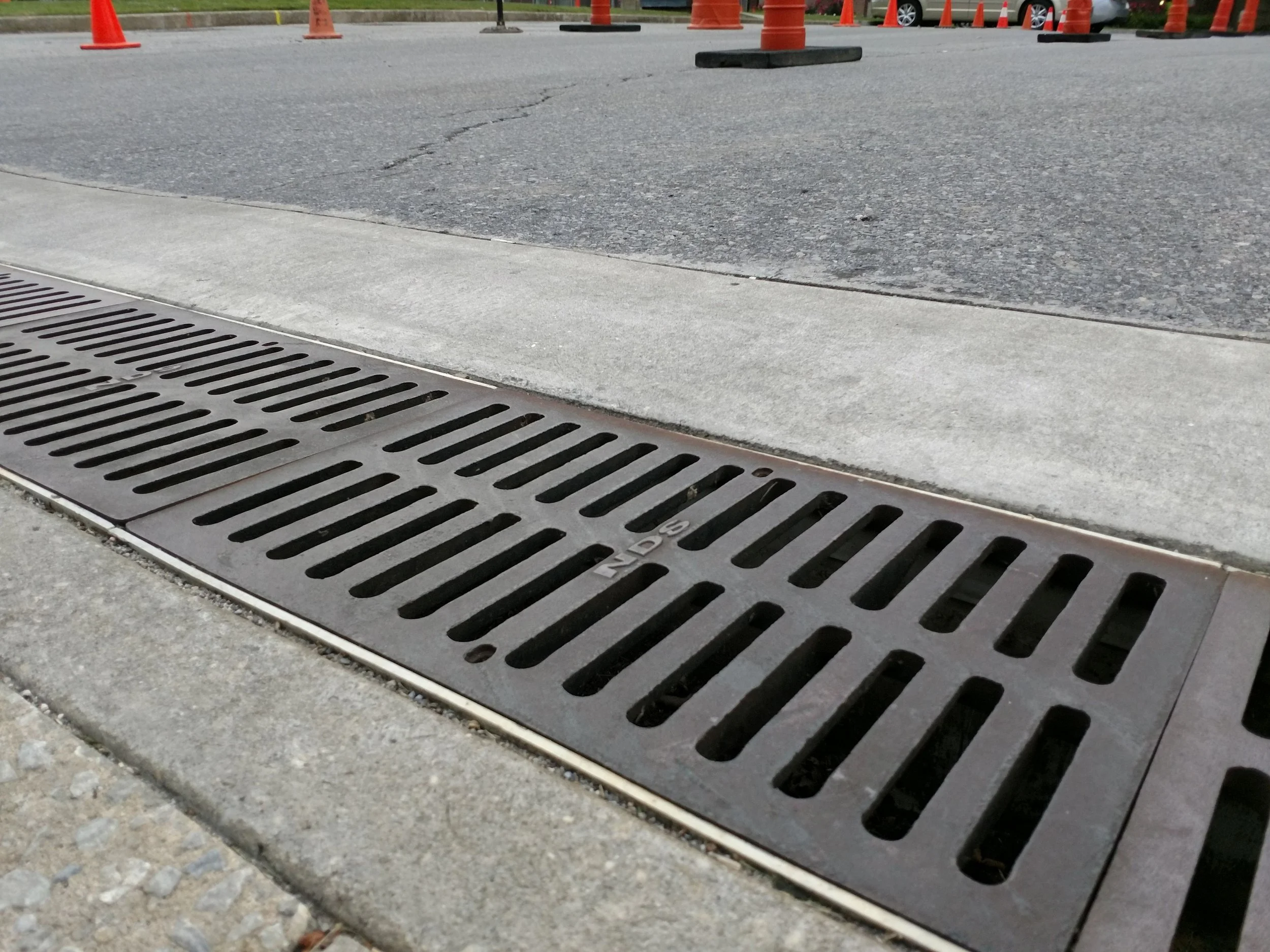 A metal storm drain grate on the side of a paved street, with orange traffic cones in the background.