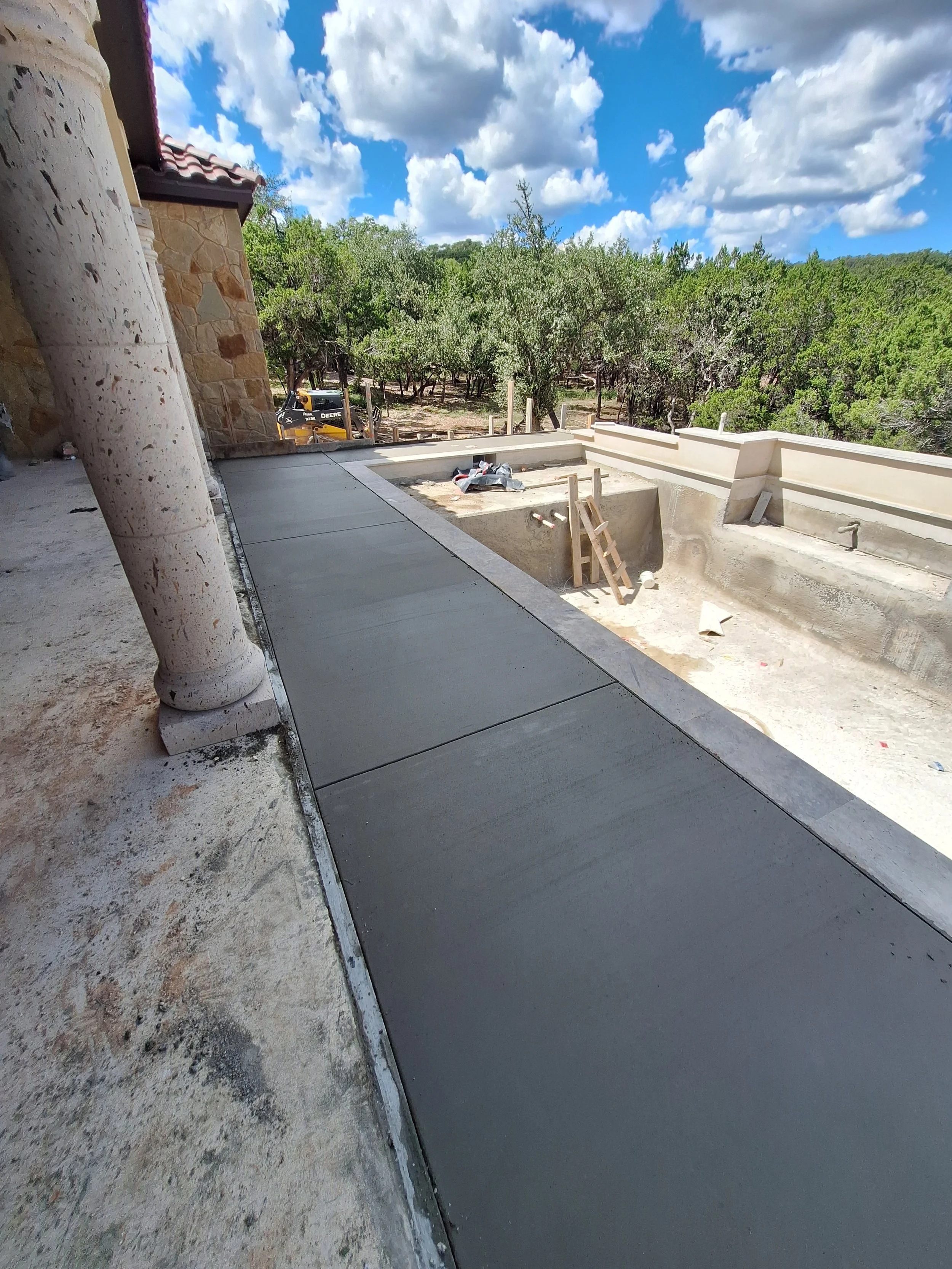 Under construction patio with new concrete sidewalk, construction materials, and scaffolding on the edge, overlooking a wooded landscape with trees and clouds.