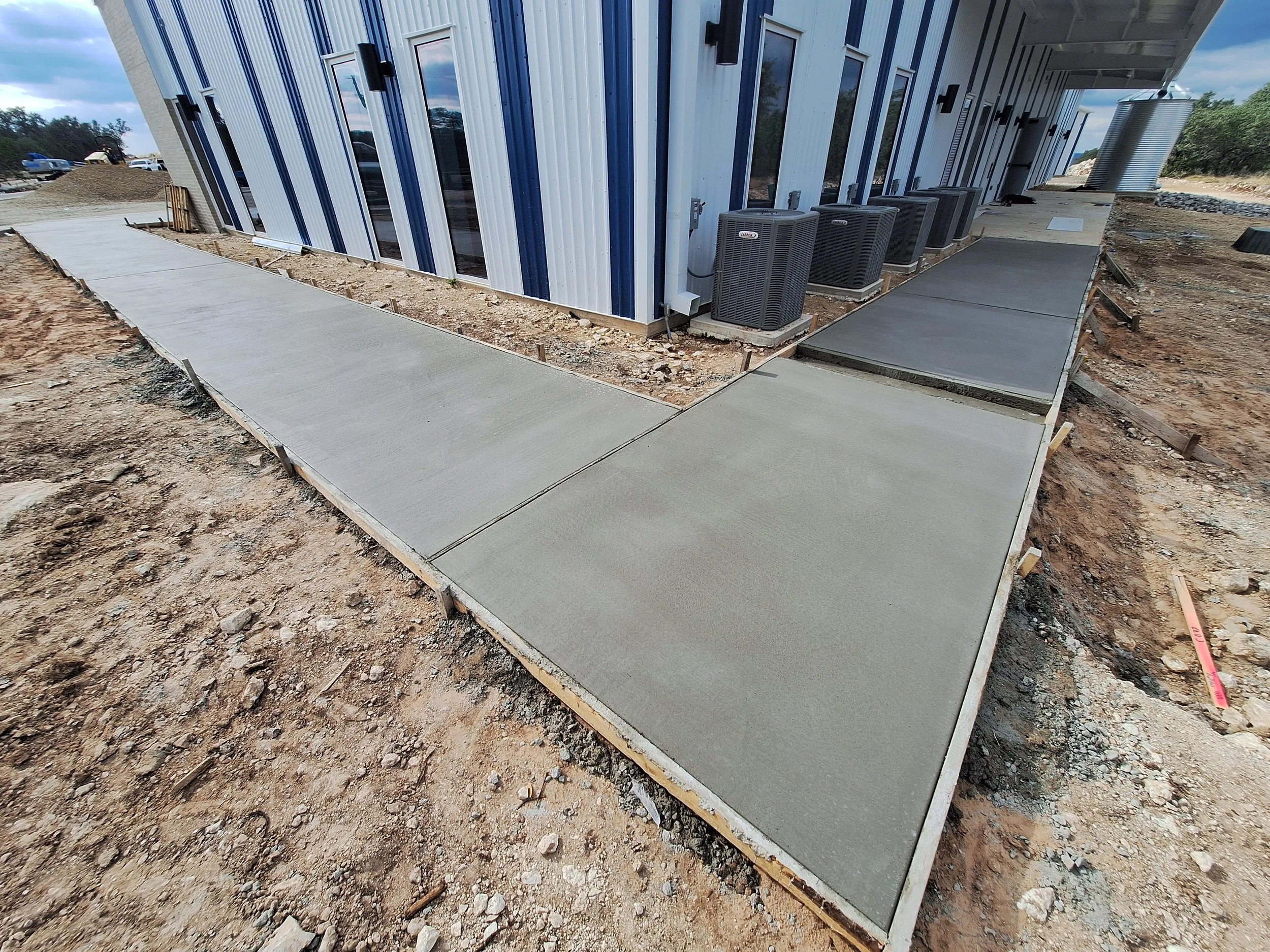 Concrete sidewalk being constructed outside a building with blue and white metal siding, air conditioning units, and a dirt ground surrounding the area.