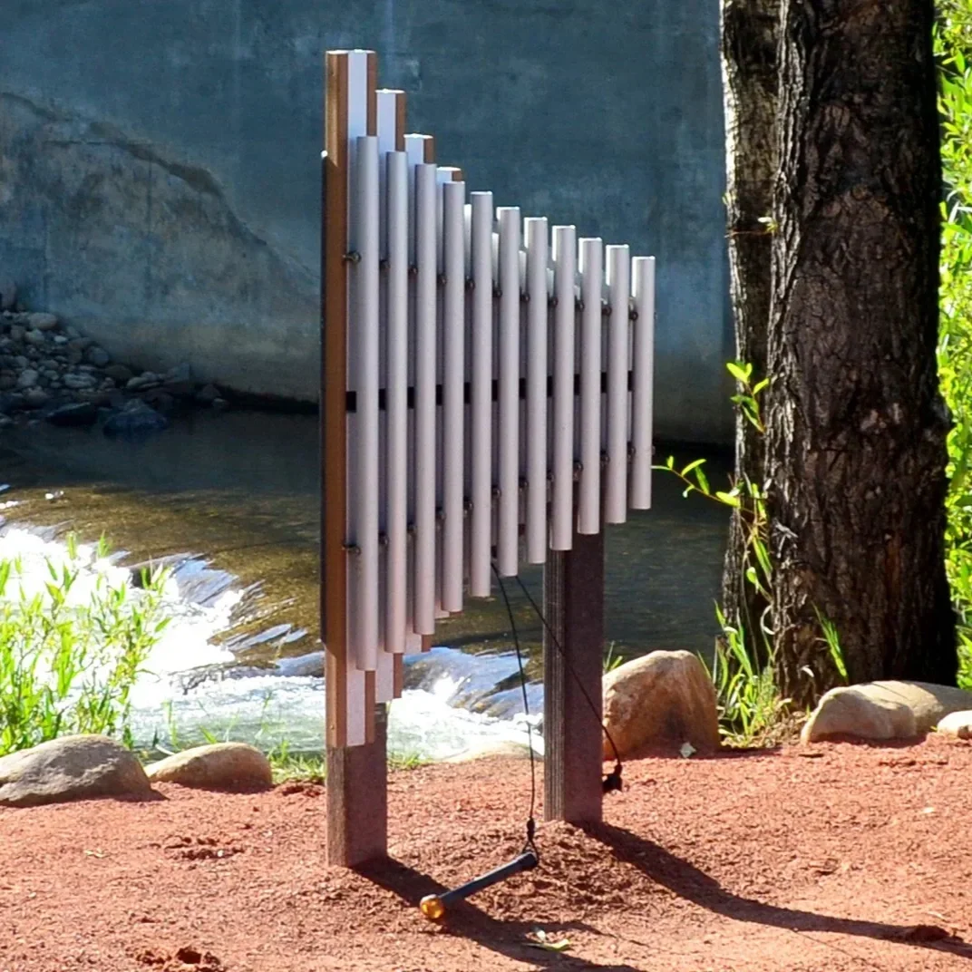 An outdoor outdoor music instrument called a wind chime made of metal tubes mounted on a wooden frame near a tree and a water stream.