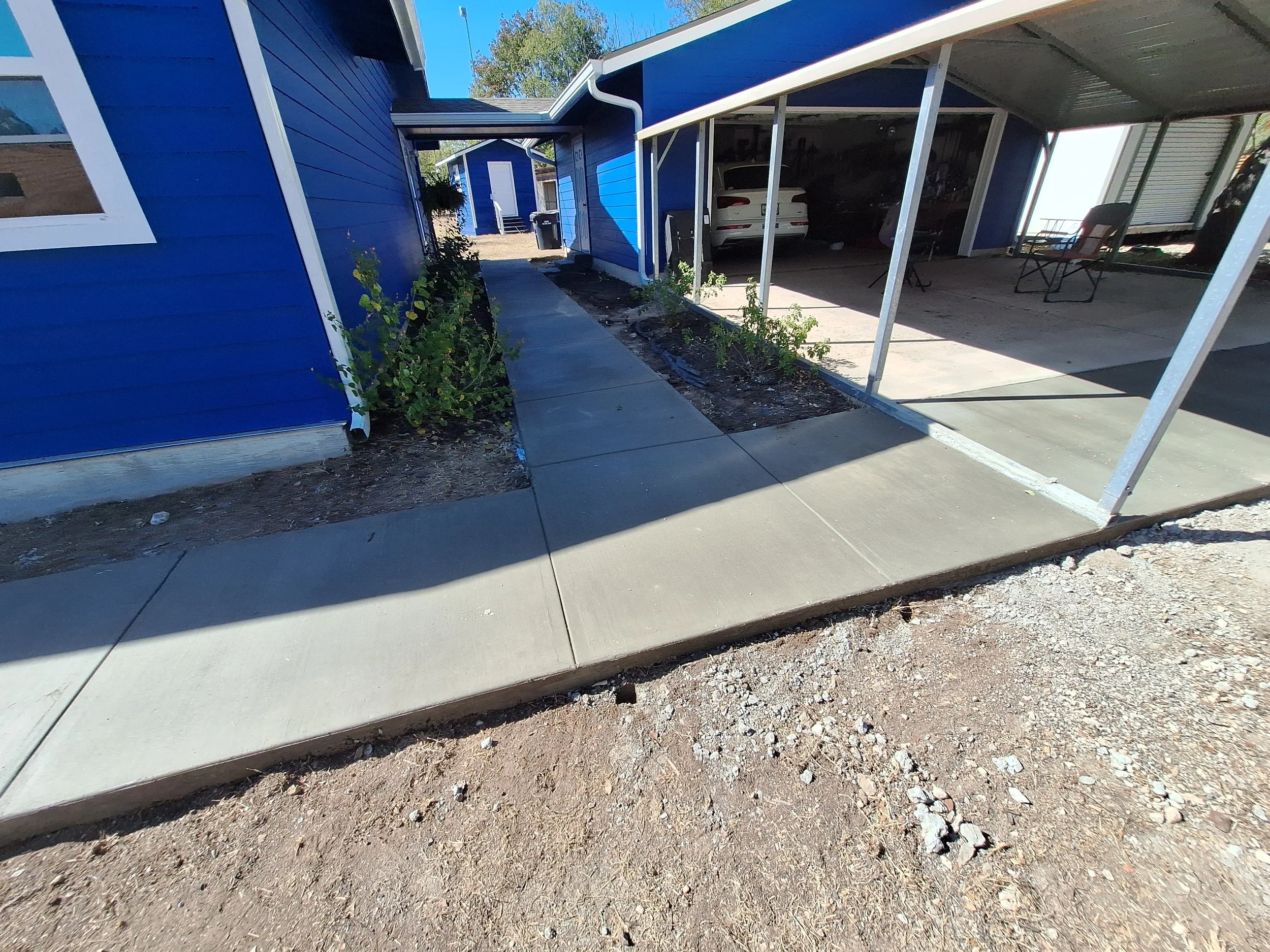 Sidewalk leading to a small blue building with a white door, with bushes on one side and a carport with parked vehicle and chairs on the other side.