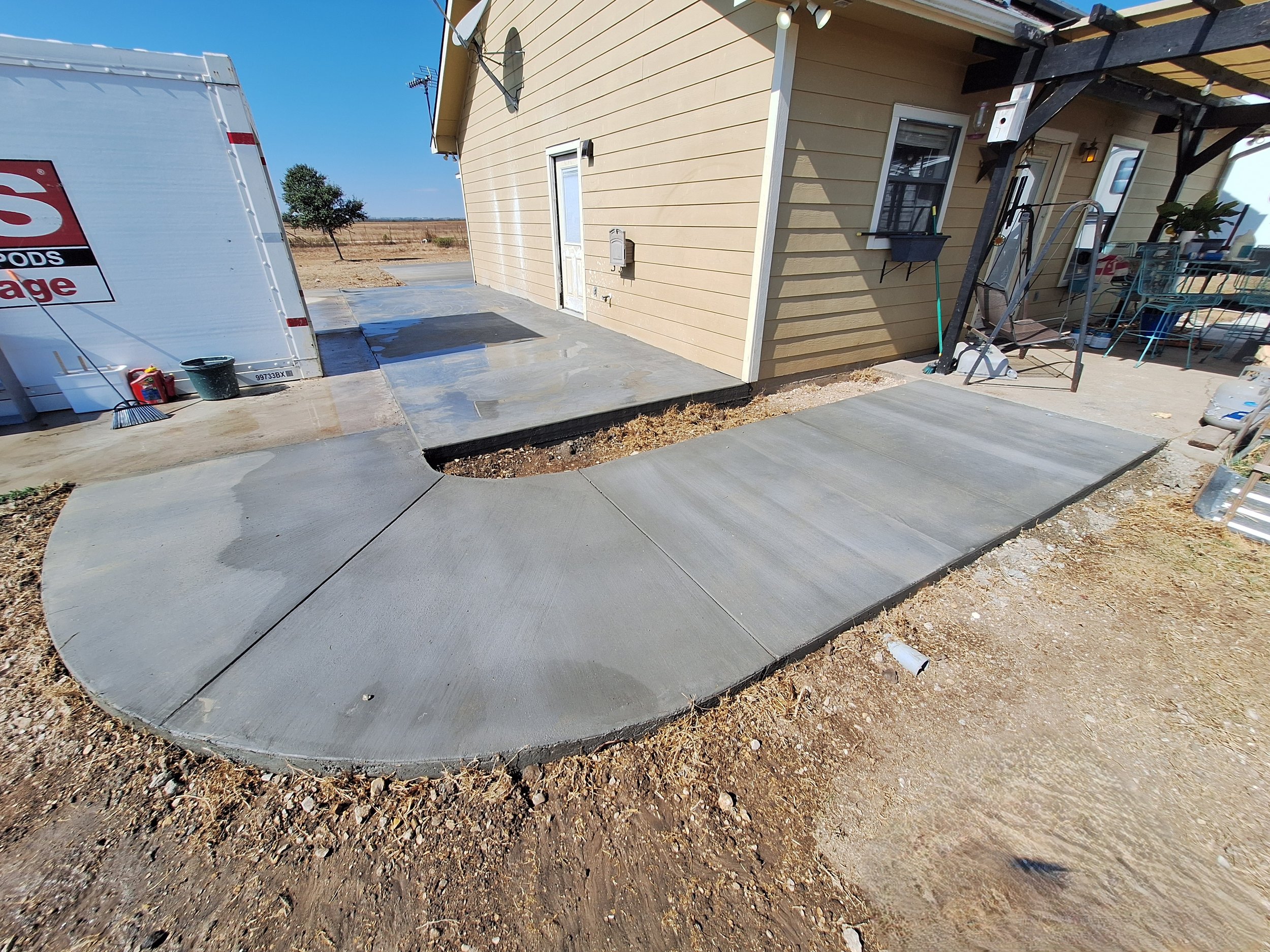 Newly concrete sidewalk with a curved section leading to a house, adjacent to a patio area with outdoor furniture and a sliding door