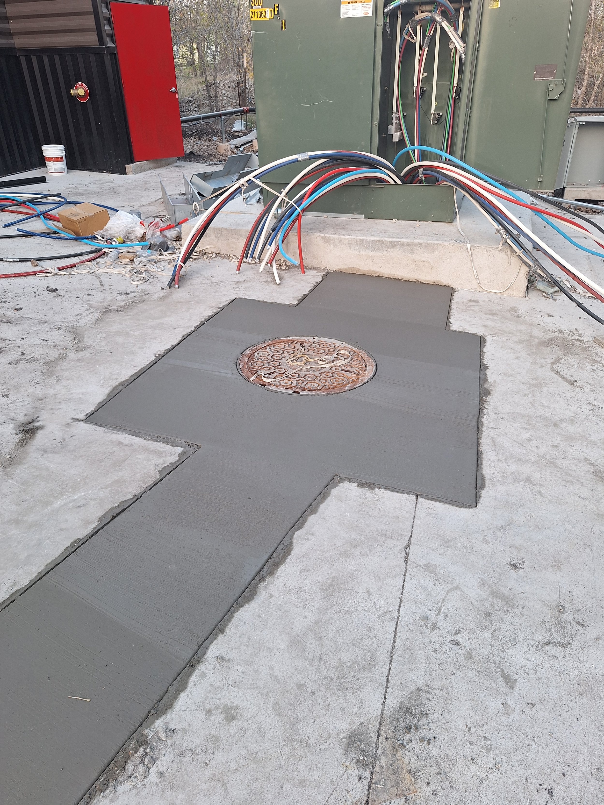 Freshly poured concrete extending around a manhole cover on a sidewalk at a construction site with exposed electrical wires and a utility box.