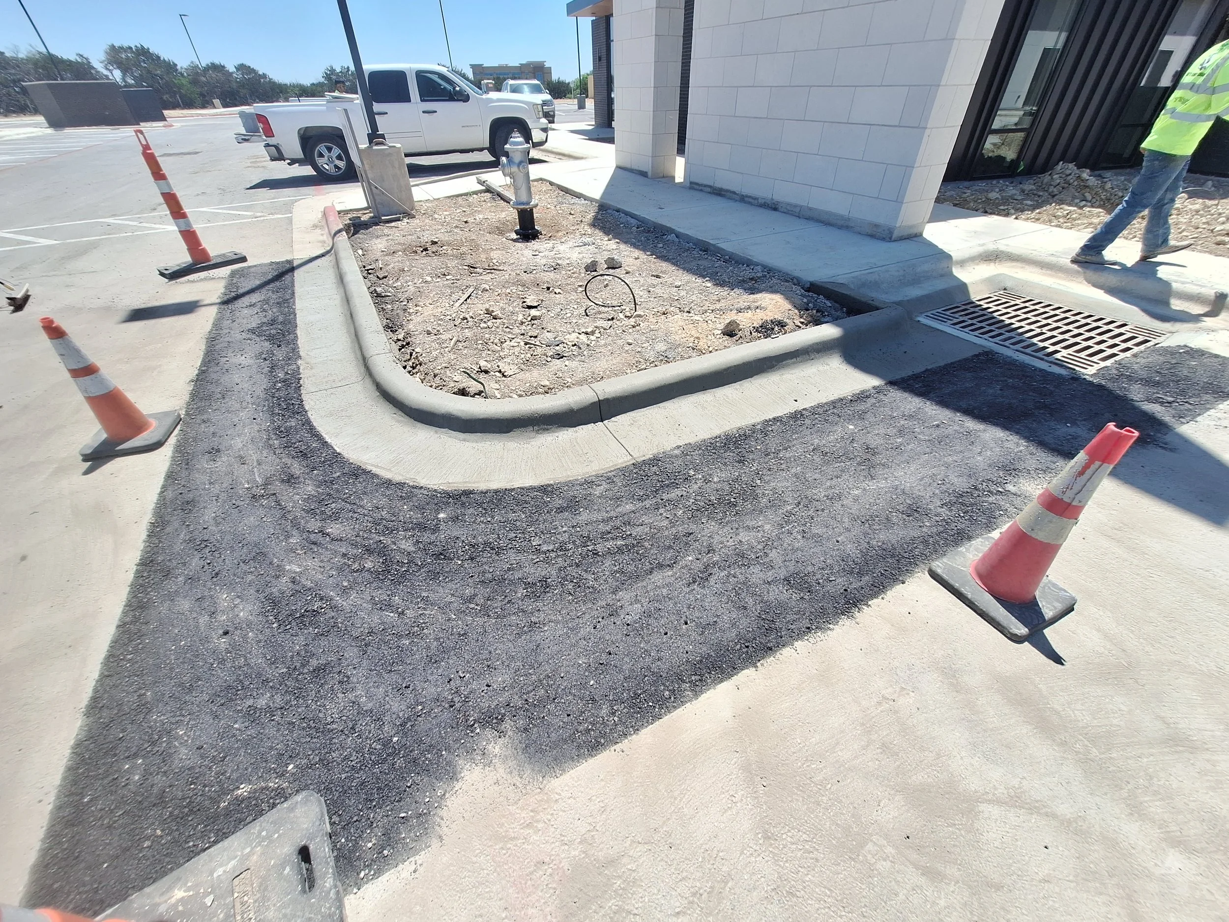 Construction site showing newly paved asphalt around a curved sidewalk, orange traffic cones, and a worker walking beside a building.