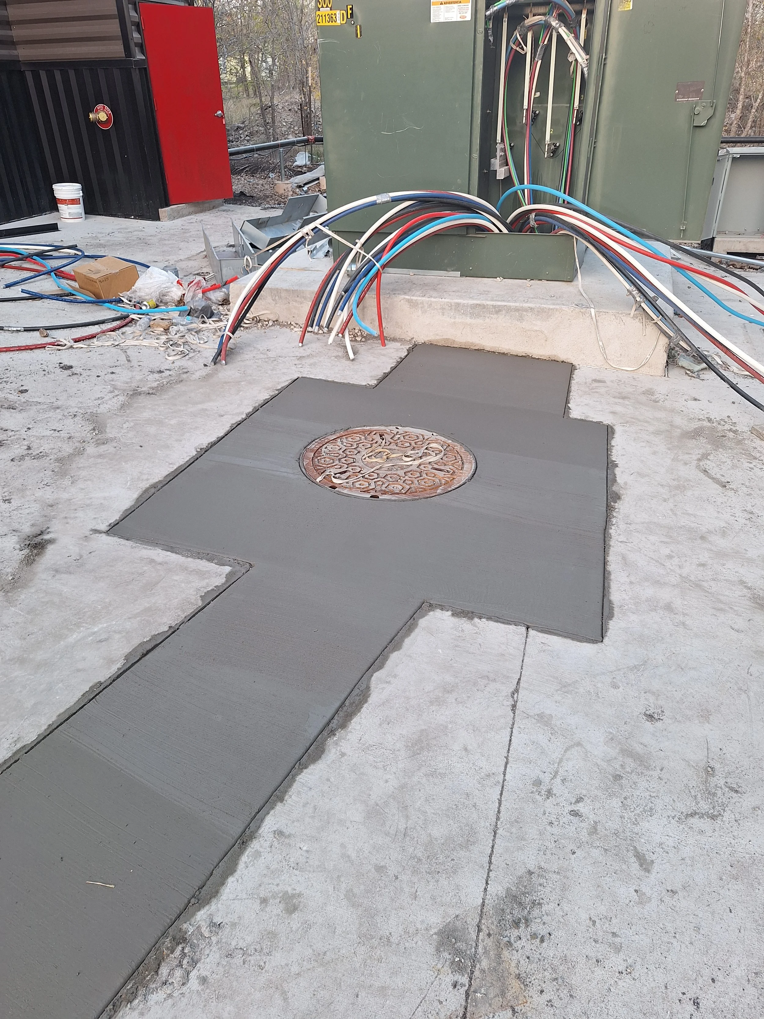 Newly poured concrete walkway with a round metal drain cover, construction wires and electrical panels in the background, and construction tools and debris surrounding the area.