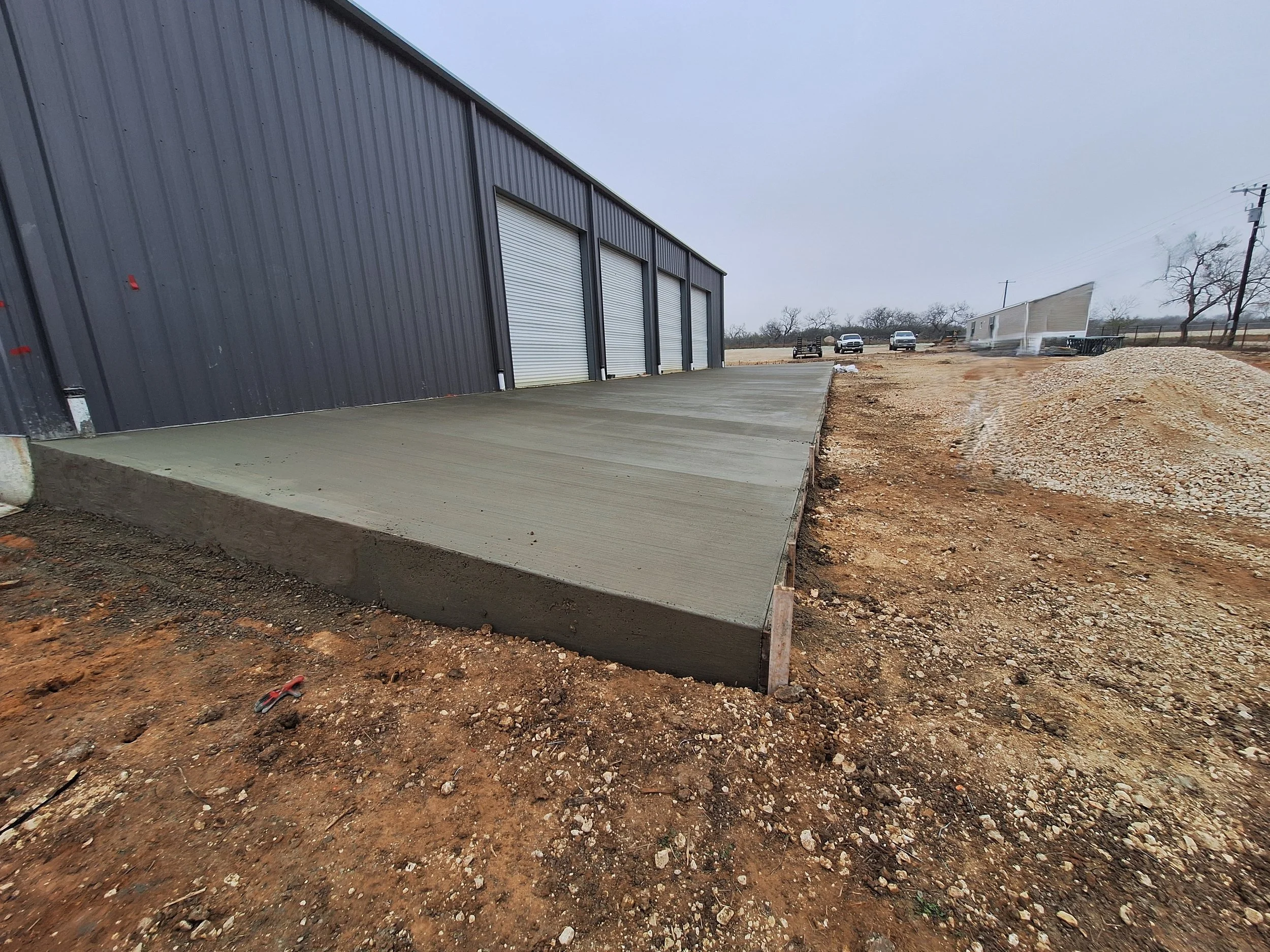 Newly poured concrete slab in front of a black metal building with three garage doors, construction site with dirt and gravel, parked cars and utility poles in the background, overcast sky.